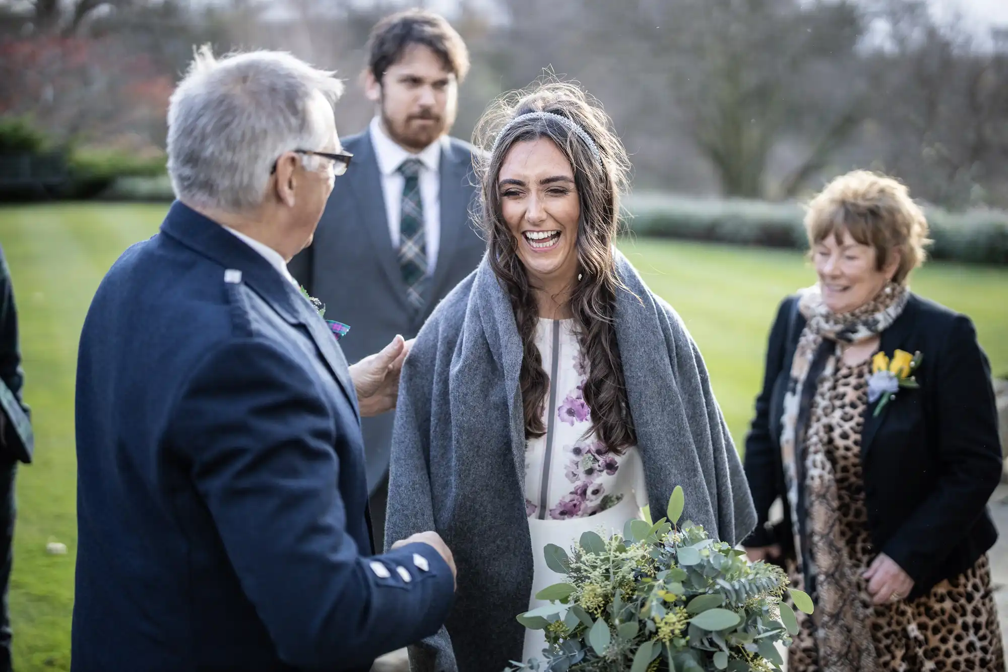 A smiling bride in a white dress with purple flowers stands outdoors, draped in a grey shawl, holding a bouquet. She is surrounded by three people—two men in suits and a woman in a leopard-print coat—on a lawn with trees in the background.