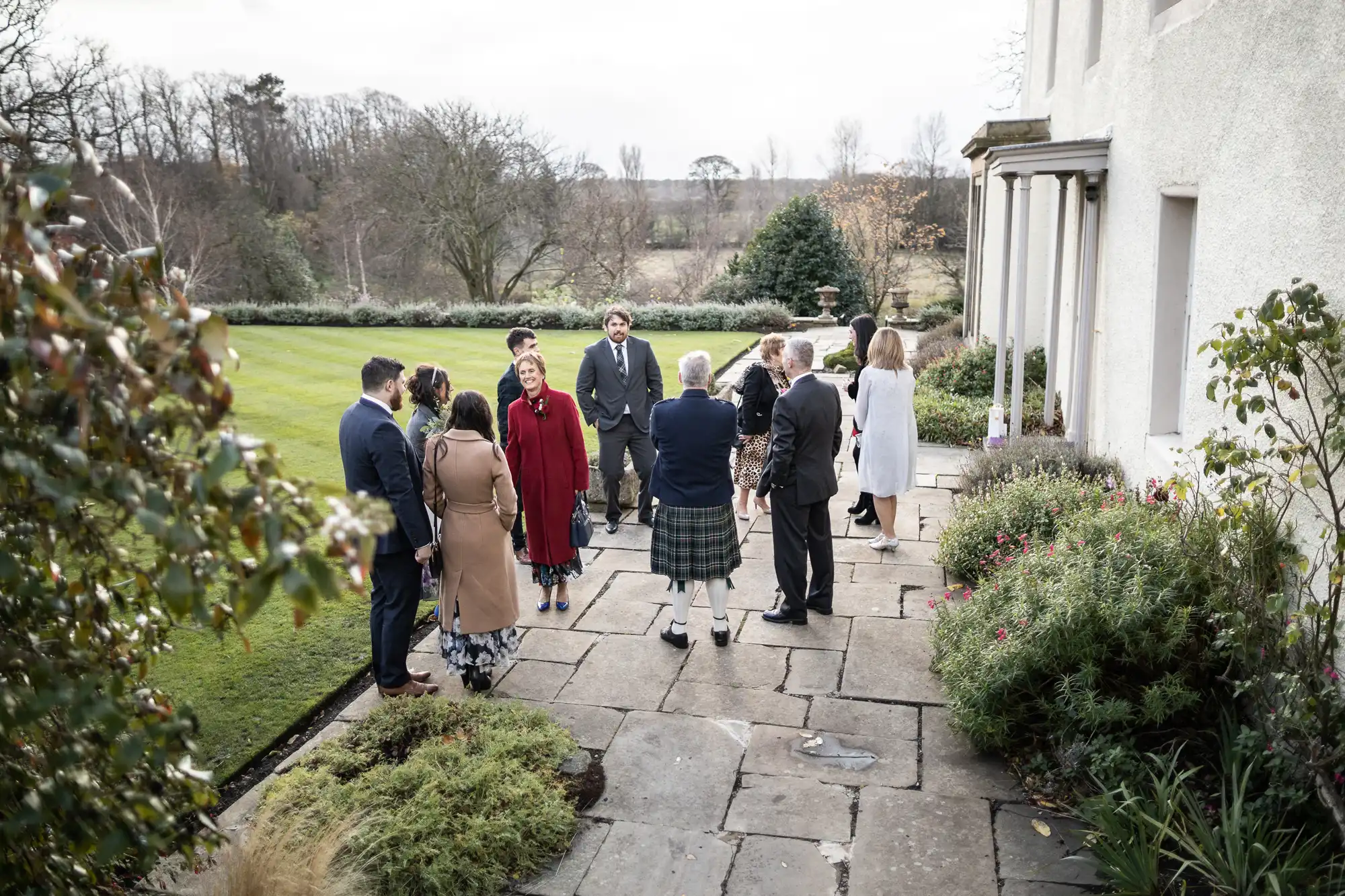 A group of people, dressed in formal and semi-formal attire, stand chatting on a large stone patio beside a white building, surrounded by greenery and gardens, with a lawn and trees in the background on an overcast day.