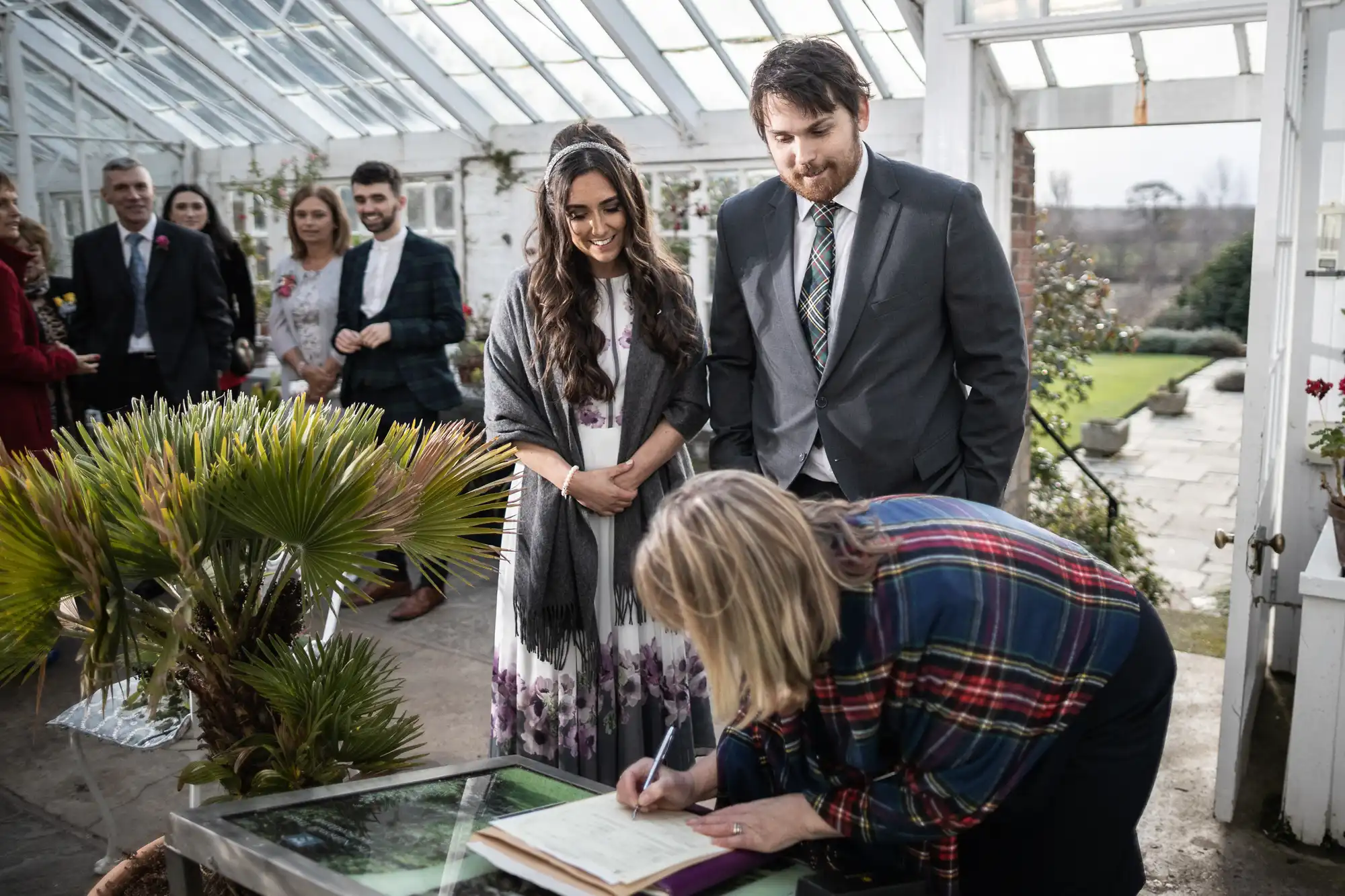 A woman signs a document on a glass table in a bright conservatory, whilst a smiling couple in formal attire stand beside her. Guests are gathered in the background, with greenery and large windows letting in natural light.
