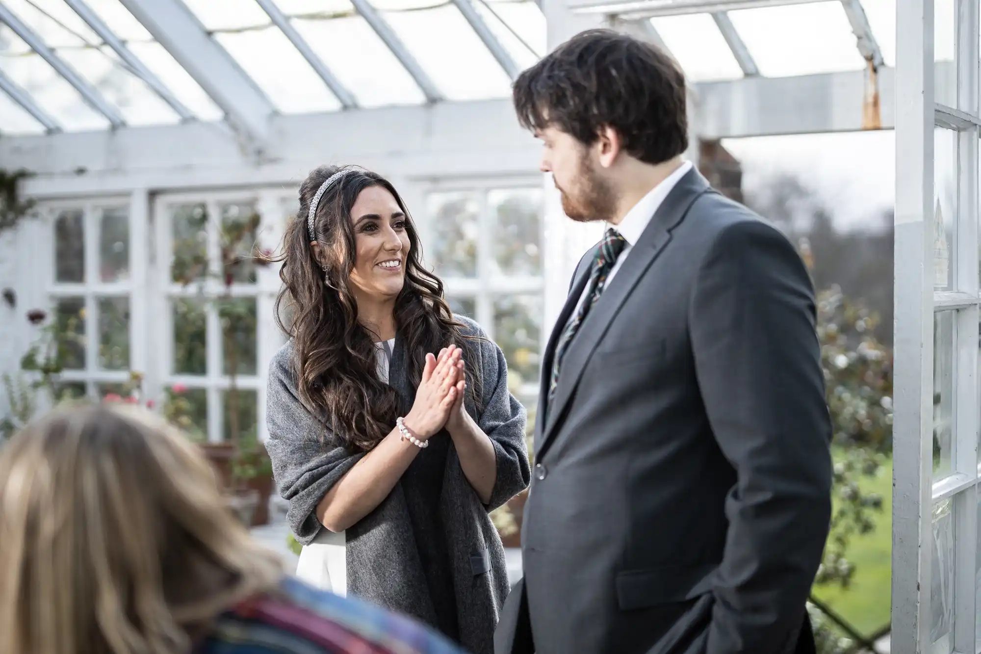 A woman with long dark hair, wearing a grey shawl and white dress, smiles with hands clasped, looking at a bearded man in a grey suit and striped tie. They stand in a bright greenhouse with large windows and plants in the background.
