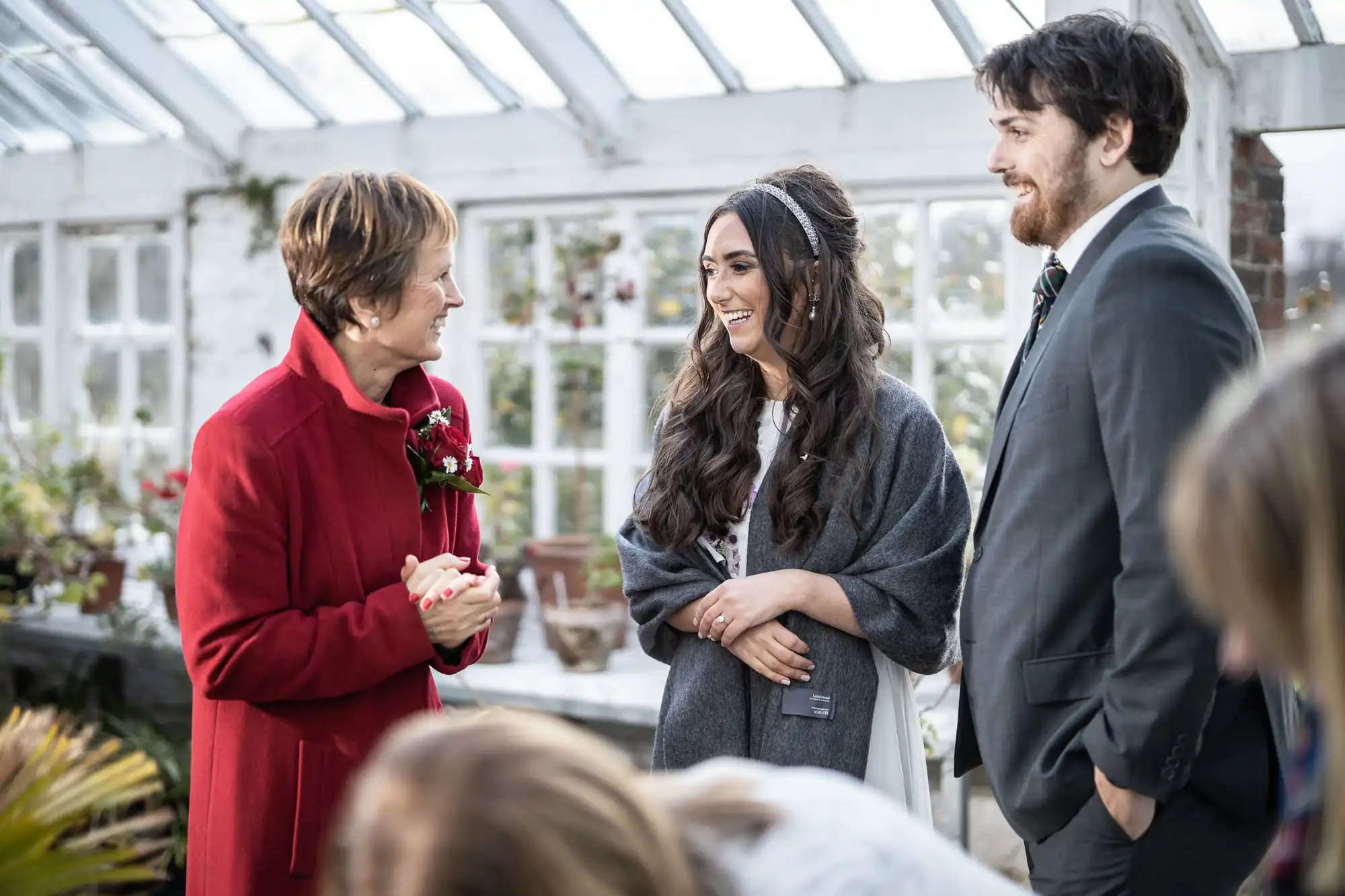 Three people stand indoors by large windows, smiling and conversing. One woman wears a red coat and buttonhole, another wears a grey shawl and white dress, and a man in a grey suit stands beside them. The background shows potted plants and sunlight.