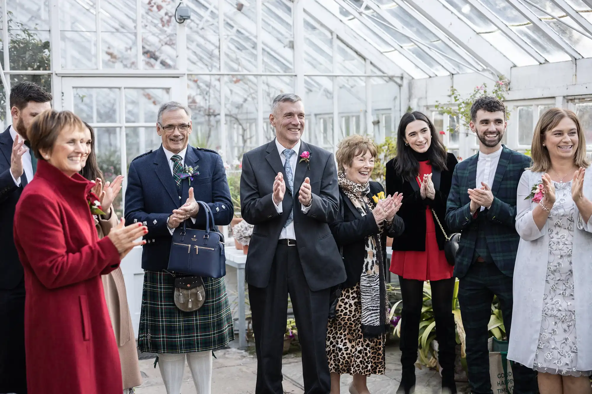 A group of people, dressed in formal and semi-formal attire, stand together in a glasshouse, smiling and clapping. Some wear suits, dresses, or patterned coats, and one man is in a kilt. The atmosphere is cheerful and festive.