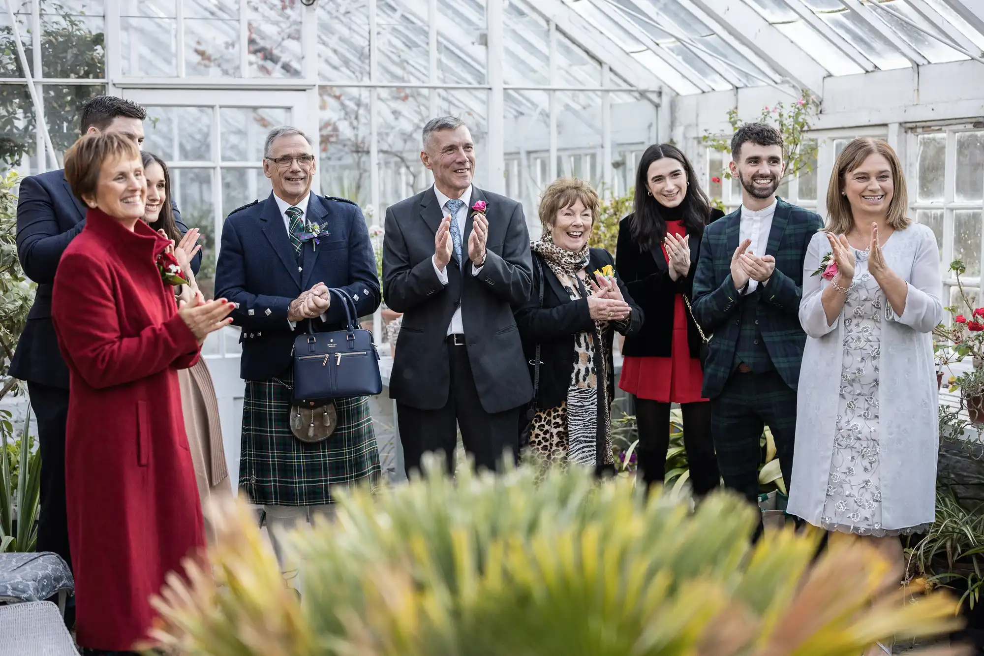 A group of nine adults stands in a sunlit greenhouse, smiling and clapping. They are dressed formally, with one man in a kilt. Lush green plants are in the foreground, and white-framed glass walls surround them. The mood is joyful and celebratory.