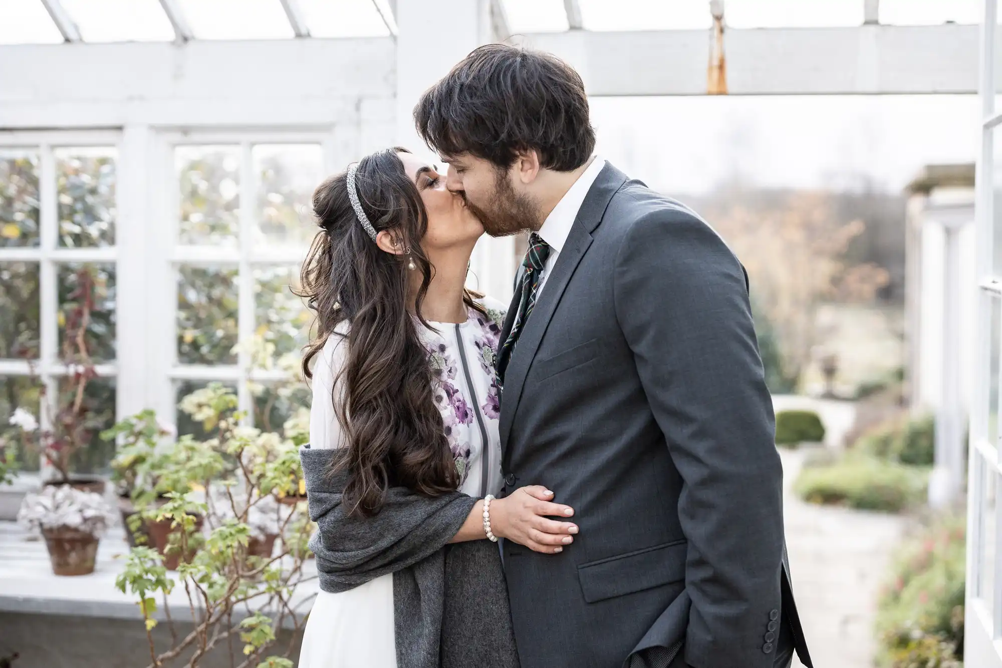 A couple snog in a bright conservatory. The woman, in a white dress with purple flowers and long dark hair, hugs the man, who wears a grey suit and tie. Plants and large windows surround them, with soft natural light filling the space.