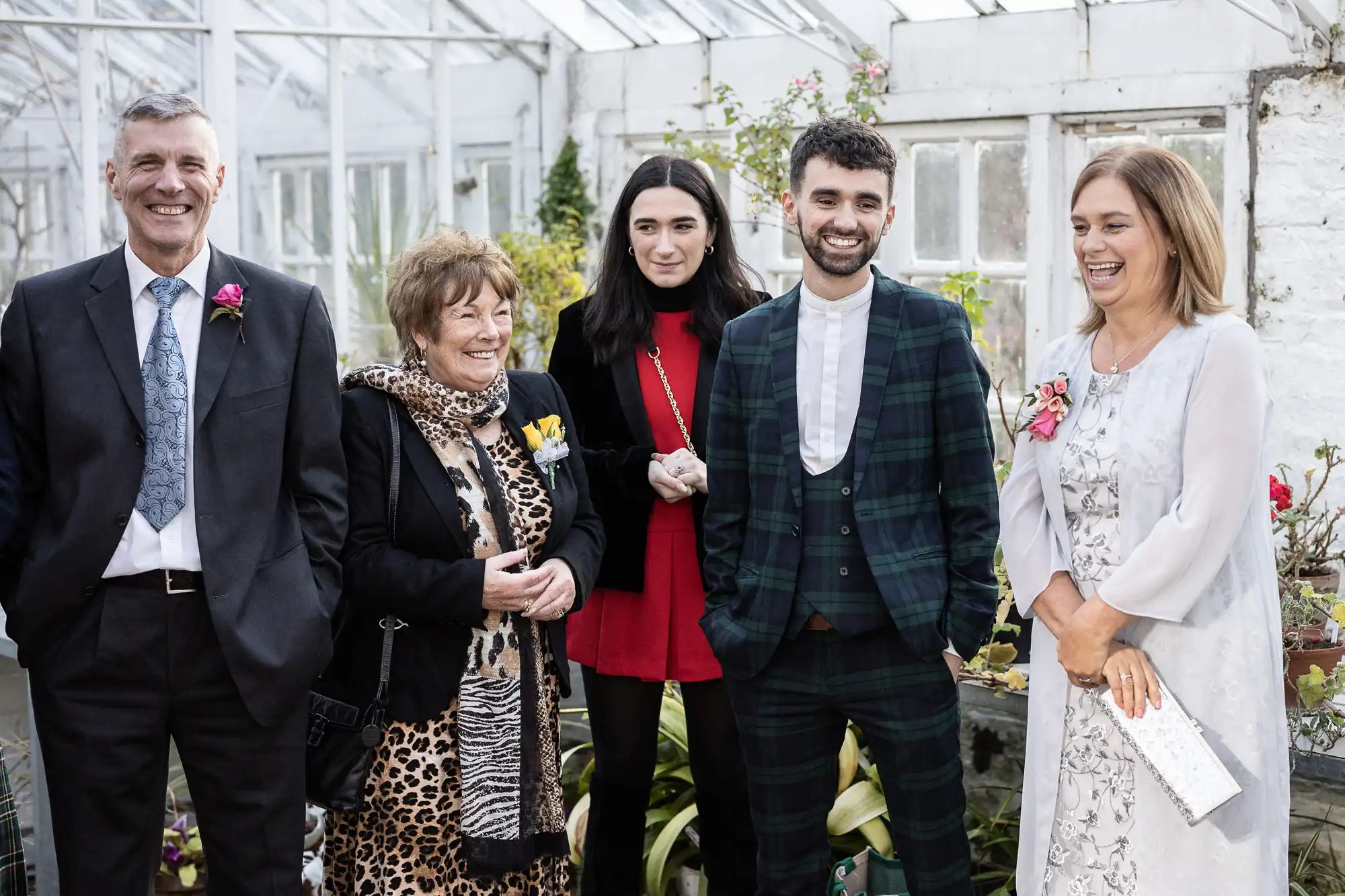 Five people stand in a greenhouse, smiling. The man on the left wears a suit and tie; the woman beside him has short hair and a leopard print scarf. The woman in the centre wears red and black; the man beside her wears a green checked suit. The woman on the right wears a pale dress.