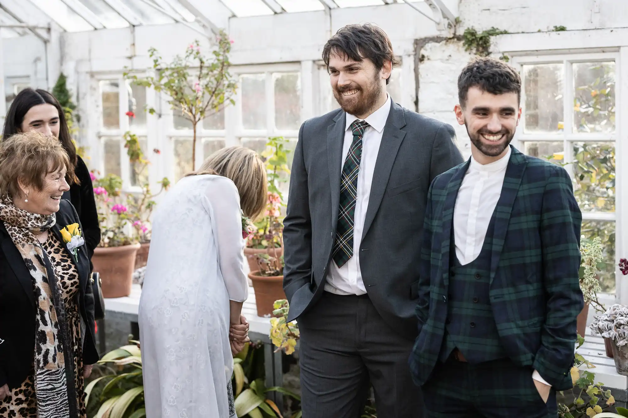 Two men in suits stand smiling in a bright glasshouse filled with potted plants. One wears a grey suit, the other a dark checked suit. Other people, including a woman in white bending forward and an elderly lady in animal print, are nearby, also smiling.