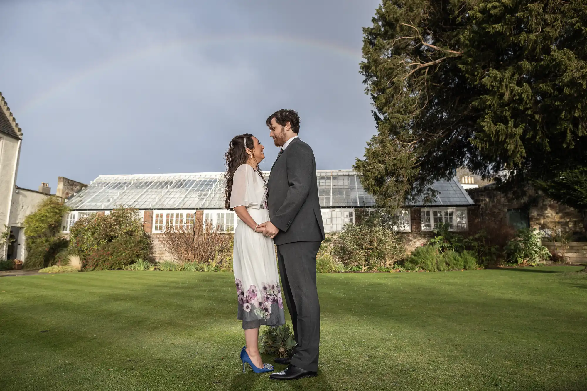 A couple stands facing each other, holding hands and smiling, on a green lawn in front of a glasshouse and garden. The woman wears a white dress with floral details and blue shoes; the man wears a grey suit. A faint rainbow arches across the cloudy sky above them.