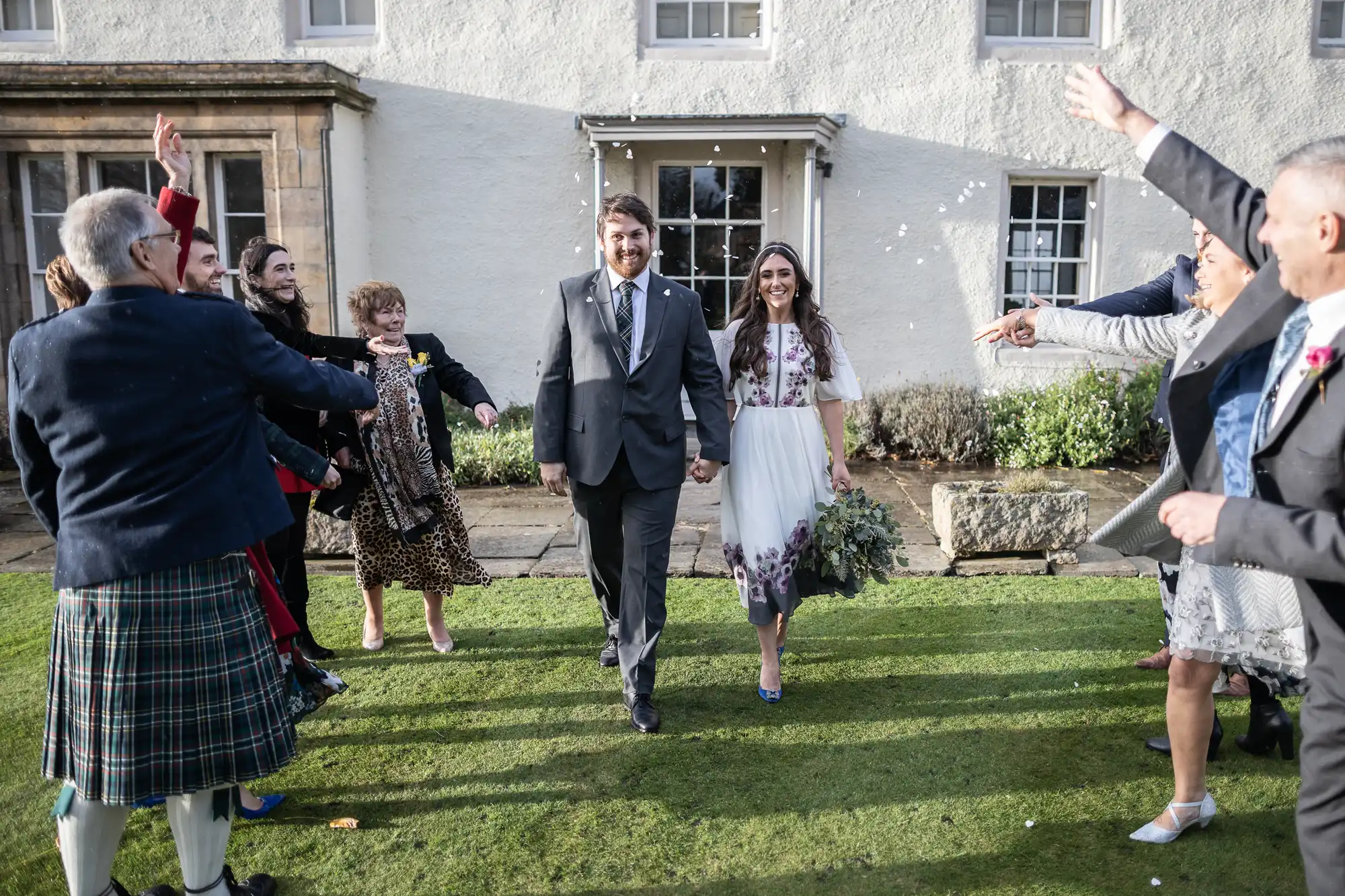 A bride and groom walk hand-in-hand on a lawn as guests shower them with confetti. The bride wears a white floral dress, and the groom a dark suit.