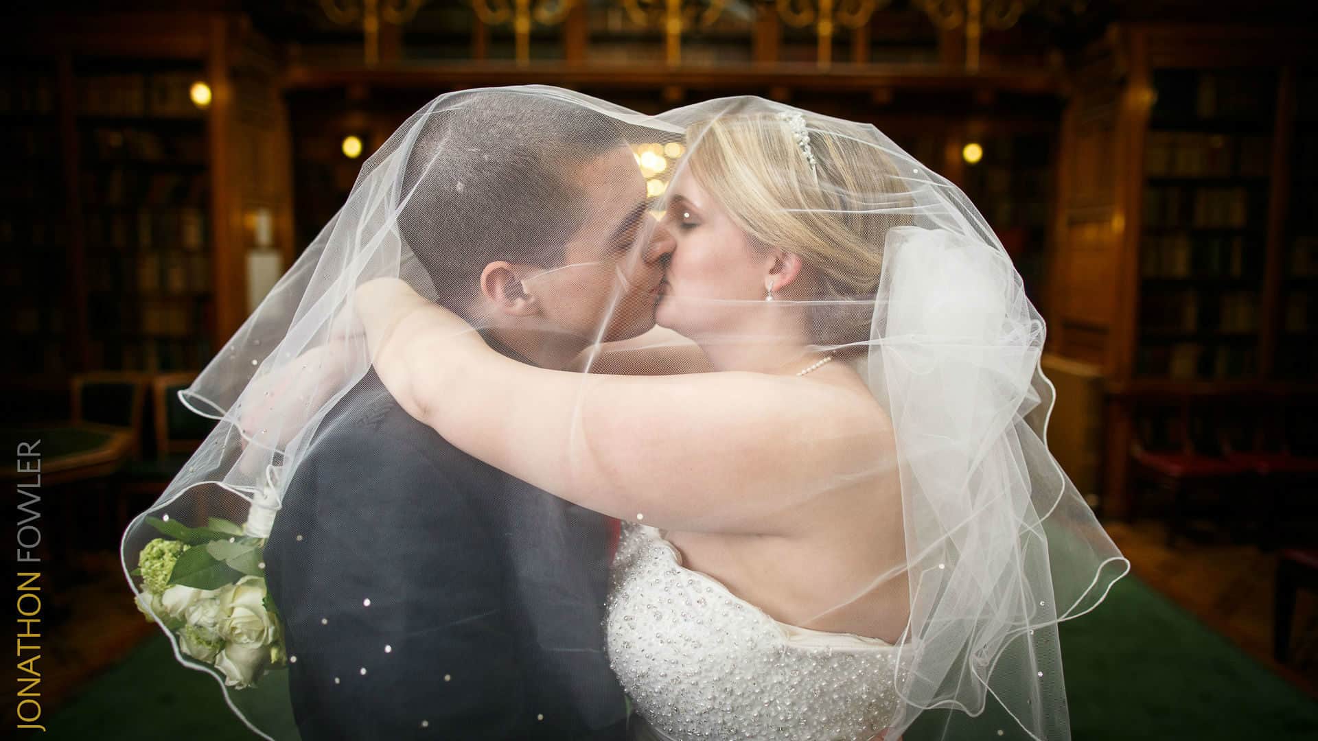 A bride and groom kiss under a translucent veil in a library with wood-paneled walls and bookshelves in the background.