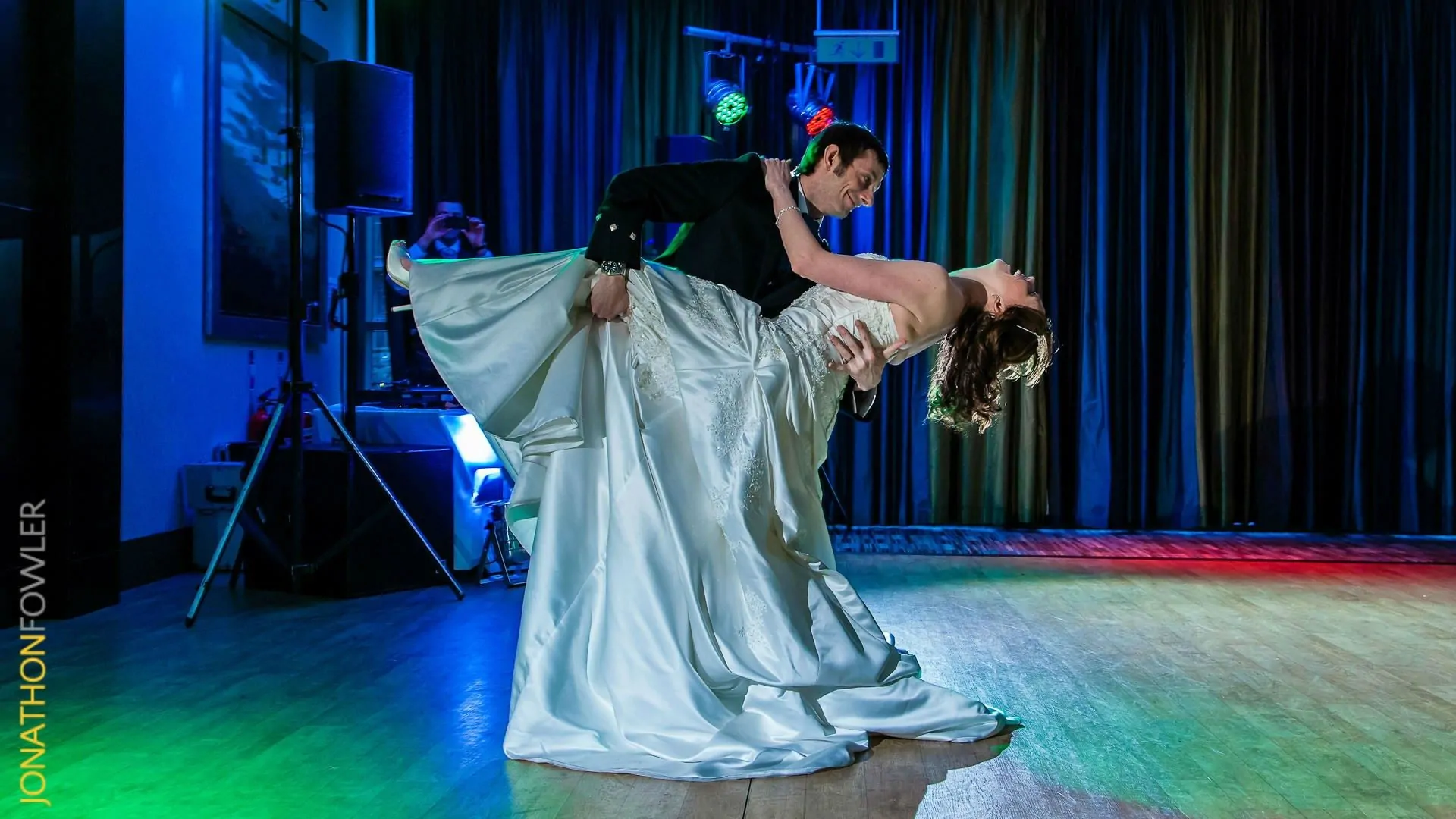 A bride and groom perform a dramatic dip dance move on a dance floor, illuminated by colorful lights.