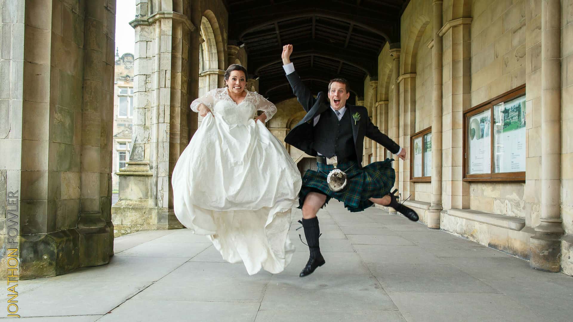 A joyful bride and groom in traditional attire jumping in the air, celebrating on a stone arcade pathway.