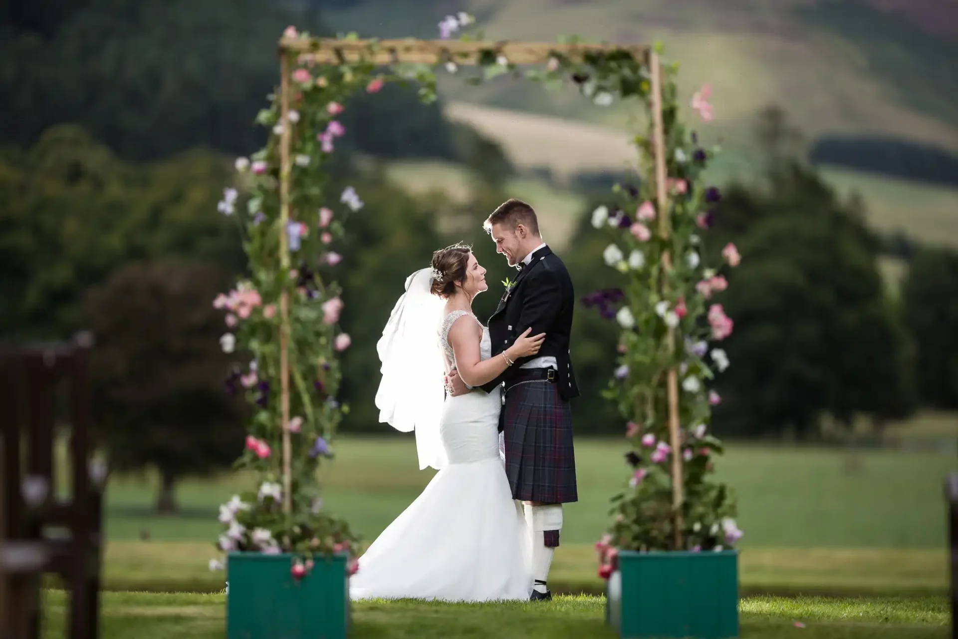 A bride in a white dress and veil and a groom in a dark jacket and traditional Scottish kilt stand smiling under a flower-adorned wooden arch outdoors at Kailzie Gardens, with green grass, trees, and rolling hills in the background.