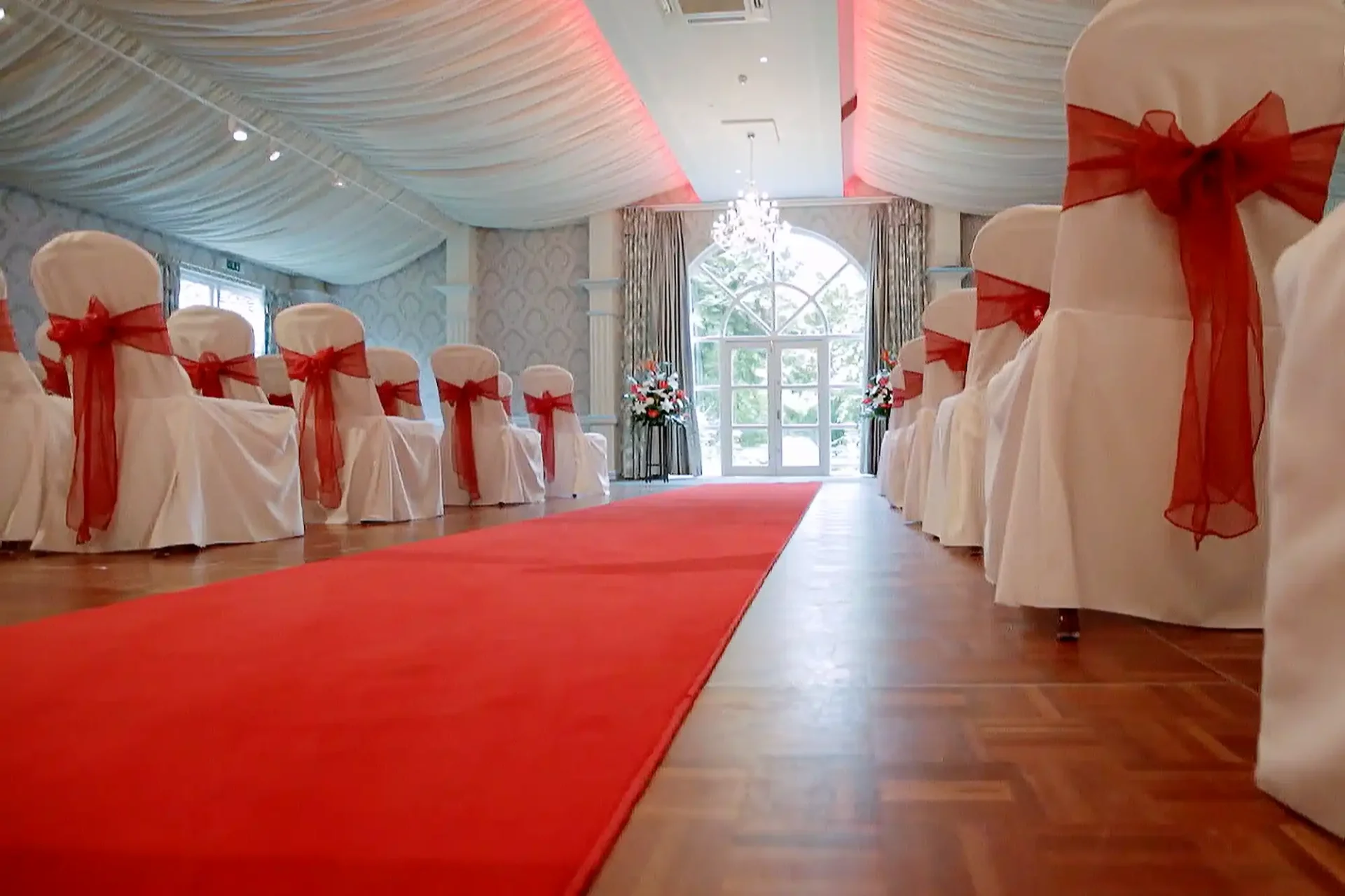 A wedding aisle with a red carpet leads to a large arched window. White chairs line the aisle, each decorated with a red organza sash. The ceiling is draped with white fabric, and floral arrangements and a chandelier are visible at the far end.