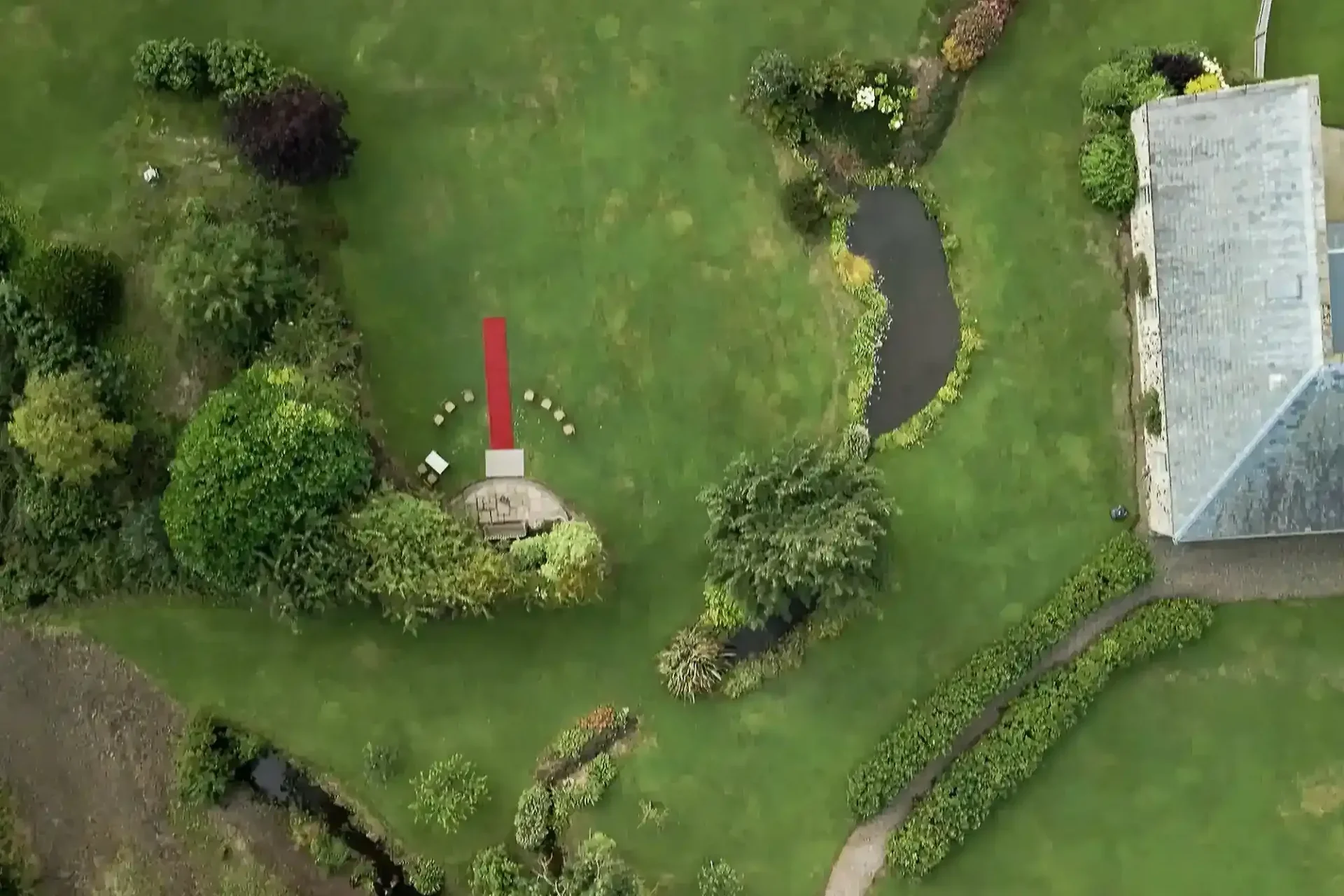 Aerial view of a lush green garden with a curved path, small pond, various shrubs and trees, and a circular patio with a red carpet leading to it. Four chairs are arranged in an arc at the patio, and a grey-roofed building is partially visible.