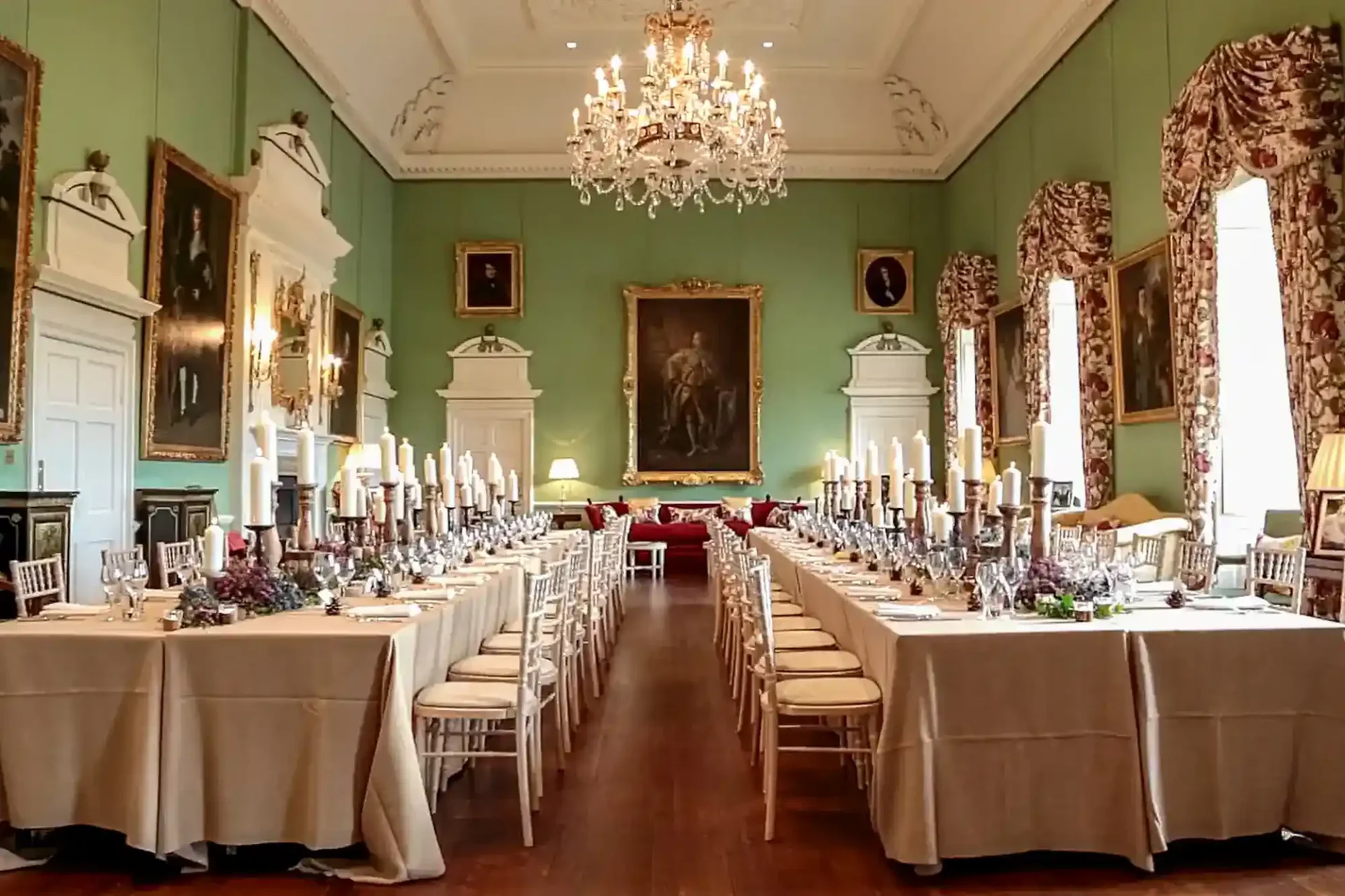 The Kinross House elegant dining hall with long tables set for a formal meal, covered in beige cloths and lined with white chairs. Chandeliers hang from the ceiling, and large classical paintings adorn the pale green walls. Tall windows are draped with patterned curtains.