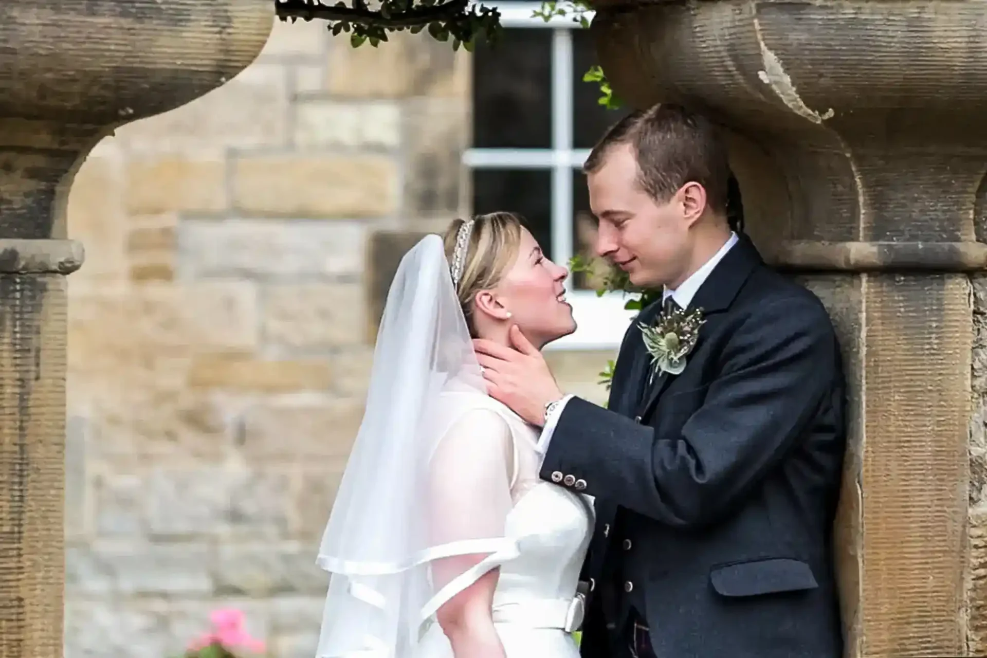 A bride and groom stand closely together under stone arches, looking into each other's eyes, dressed in wedding attire at Kirknewton House Stables.