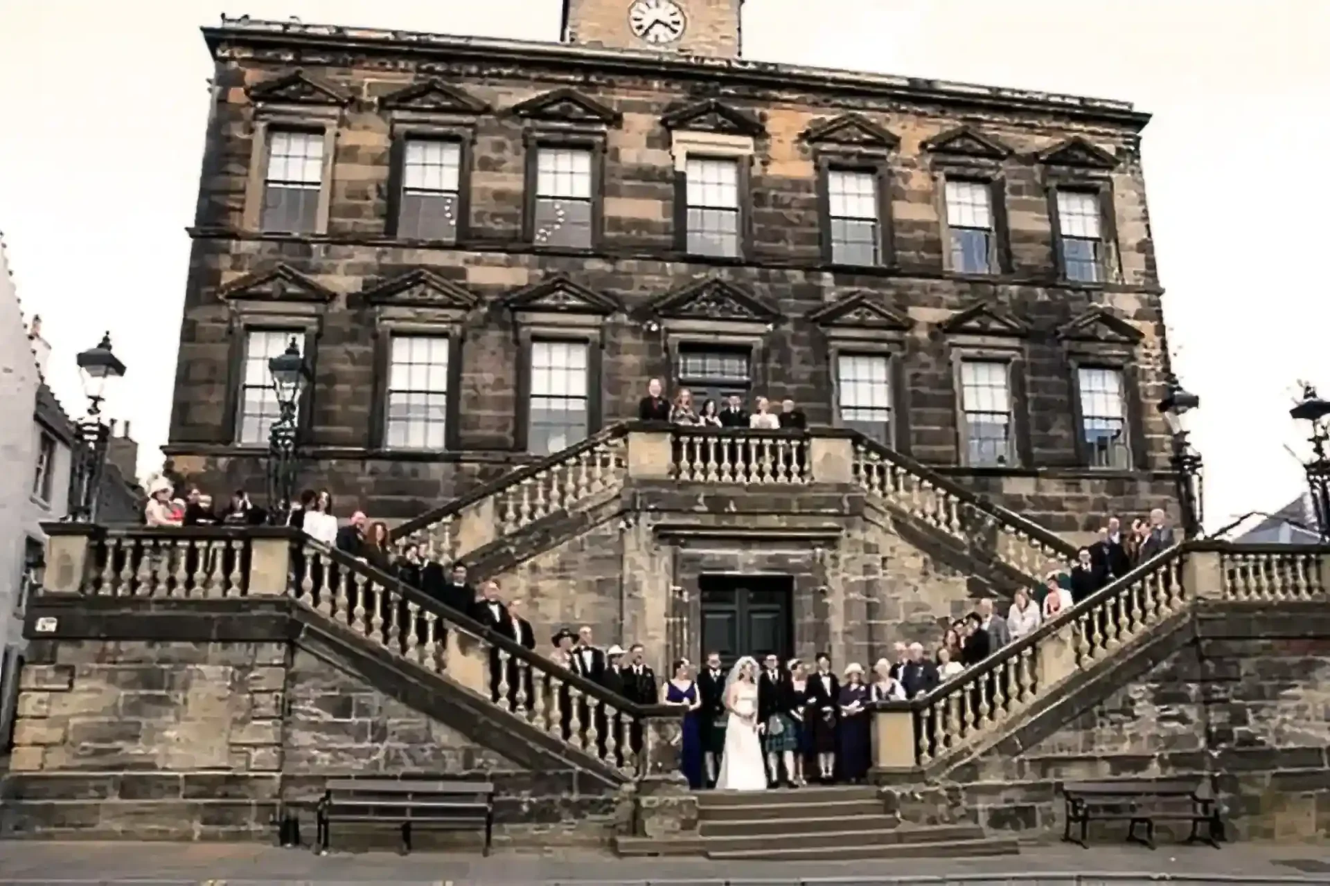 A large group of people pose for a photo on the steps of a historic stone building, Linlithgow Burgh Halls, with a clock above the entrance.
