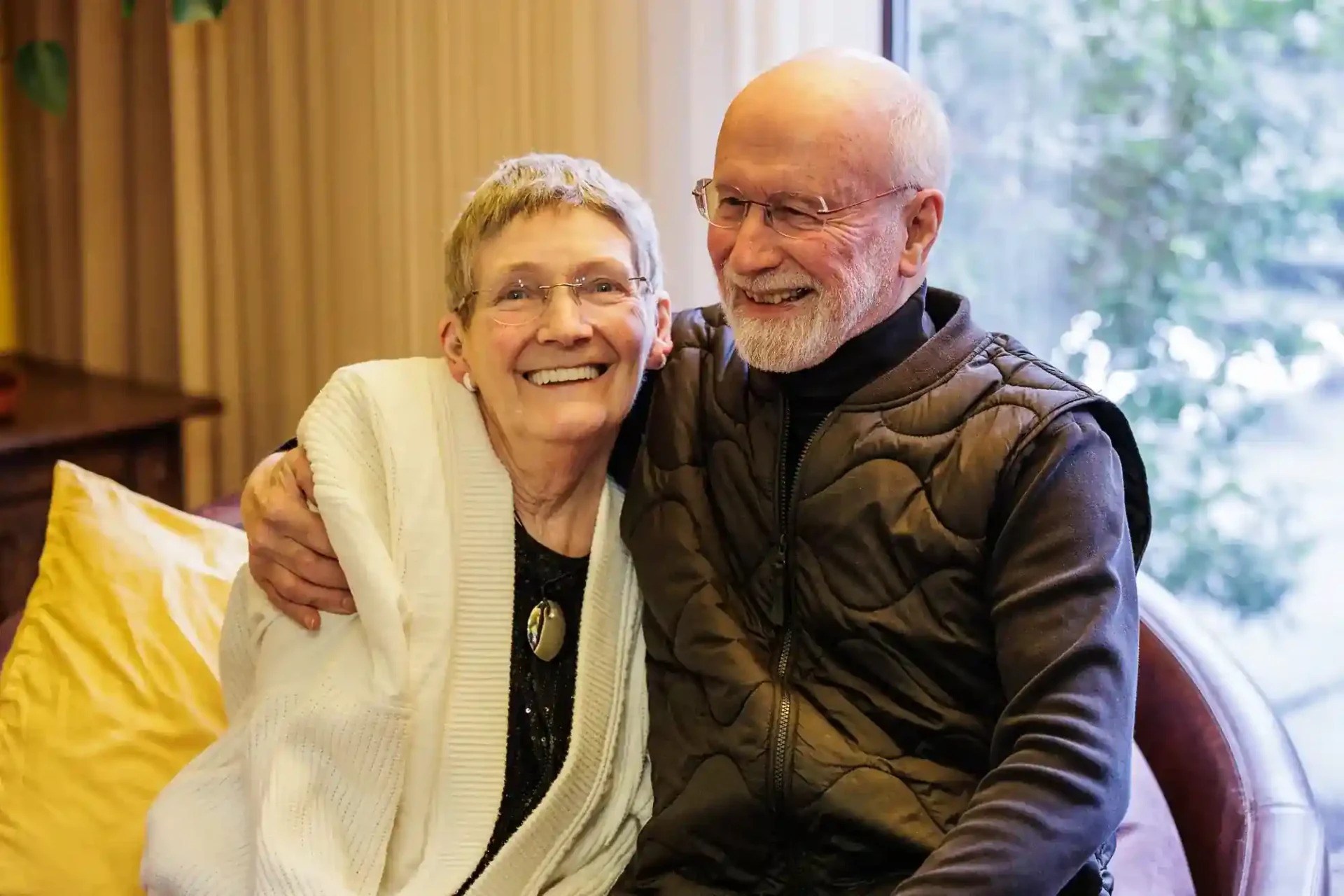An older woman and man sit closely together, smiling and laughing. The man has his arm round the woman. They appear happy and relaxed in a cosy indoor setting with a large window and soft, natural light in the background.