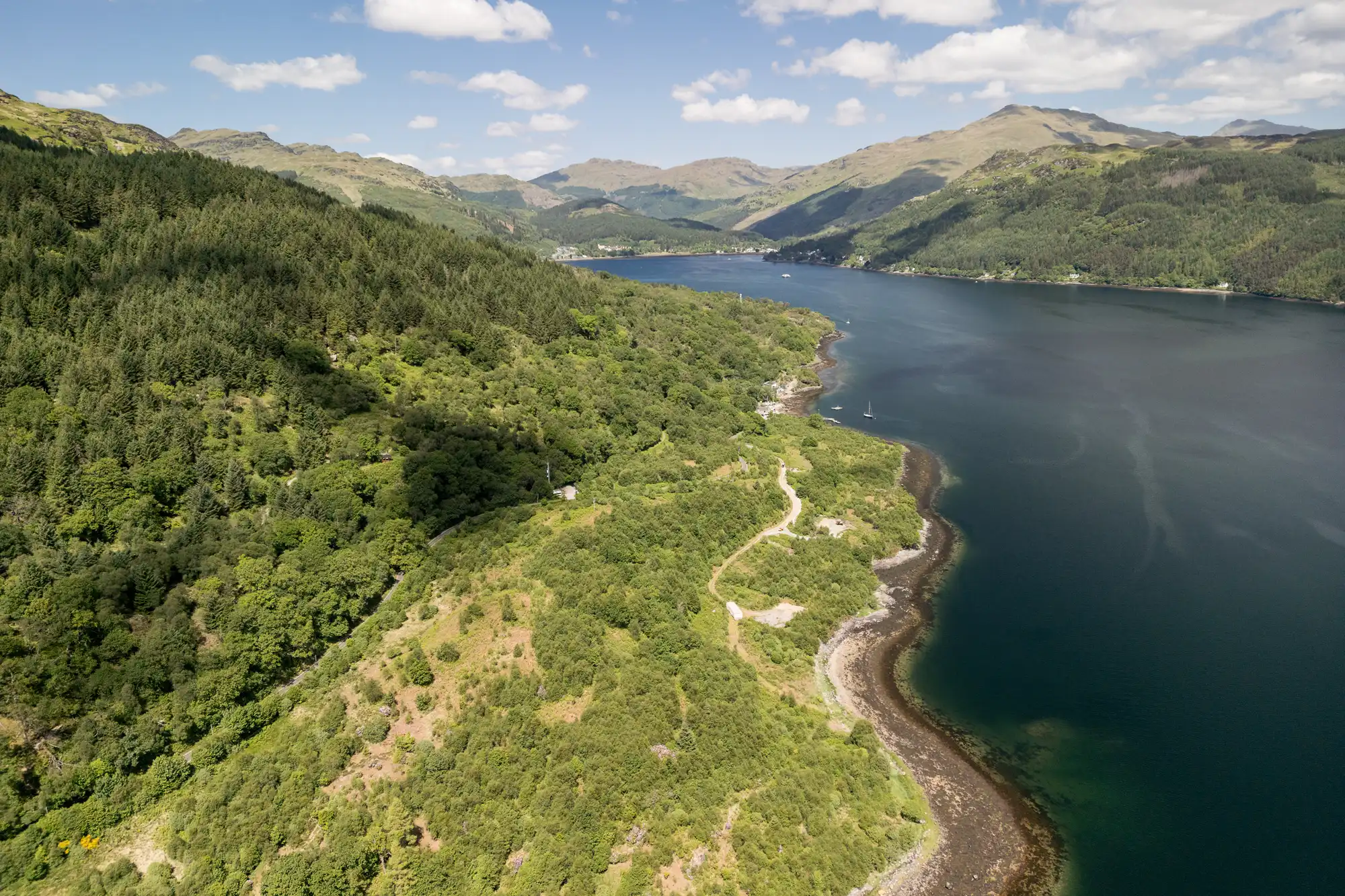 Aerial view of a lush green landscape with dense forests, winding roads, and a rocky shoreline beside a deep blue lake. Rolling hills and mountains stretch into the distance under a blue sky dotted with scattered white clouds.