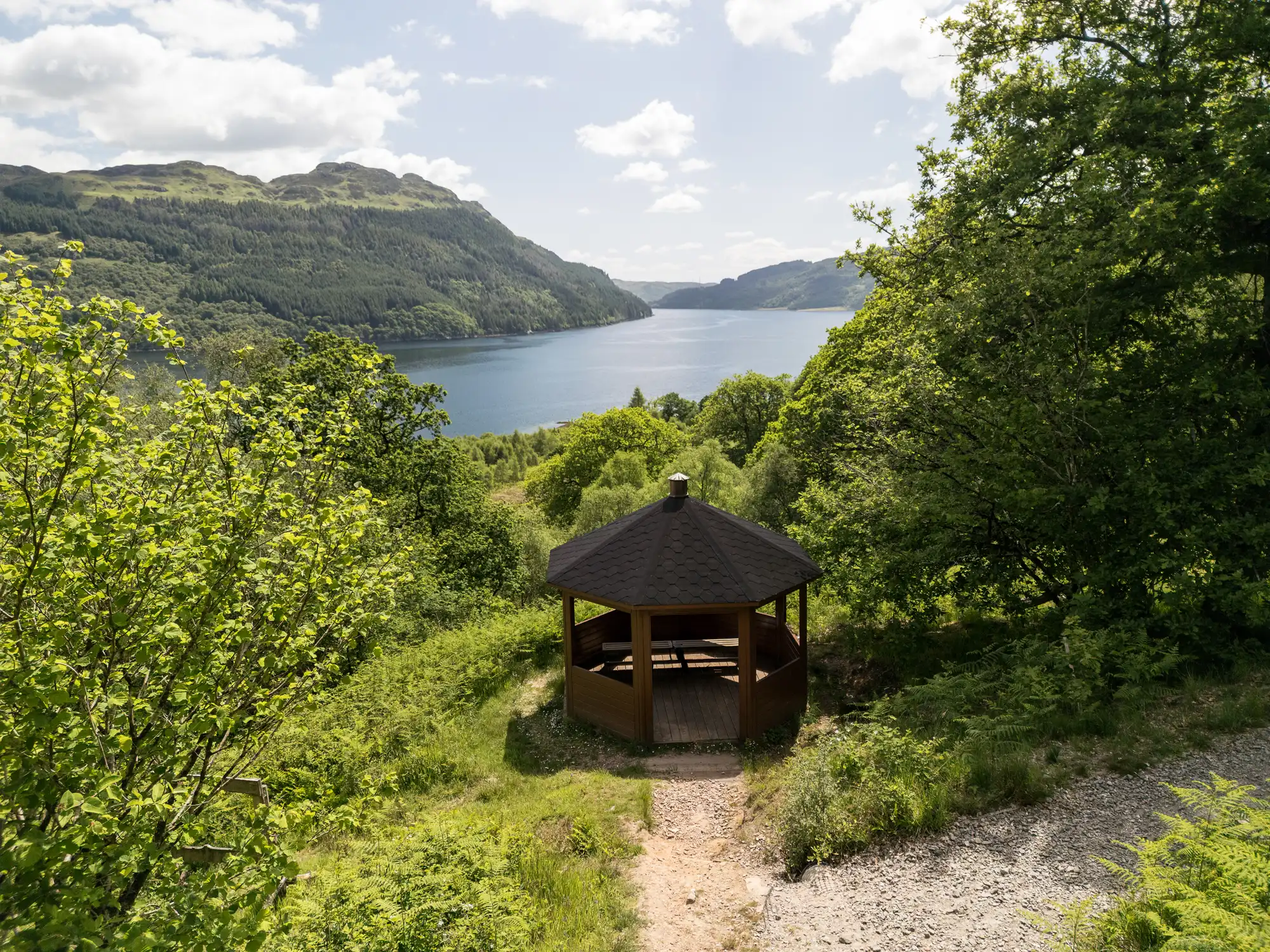 A hexagonal wooden gazebo with a dark roof sits on a hilltop, surrounded by lush green trees, overlooking a calm lake and forested mountains under a partly cloudy sky. A narrow dirt path leads to the gazebo.