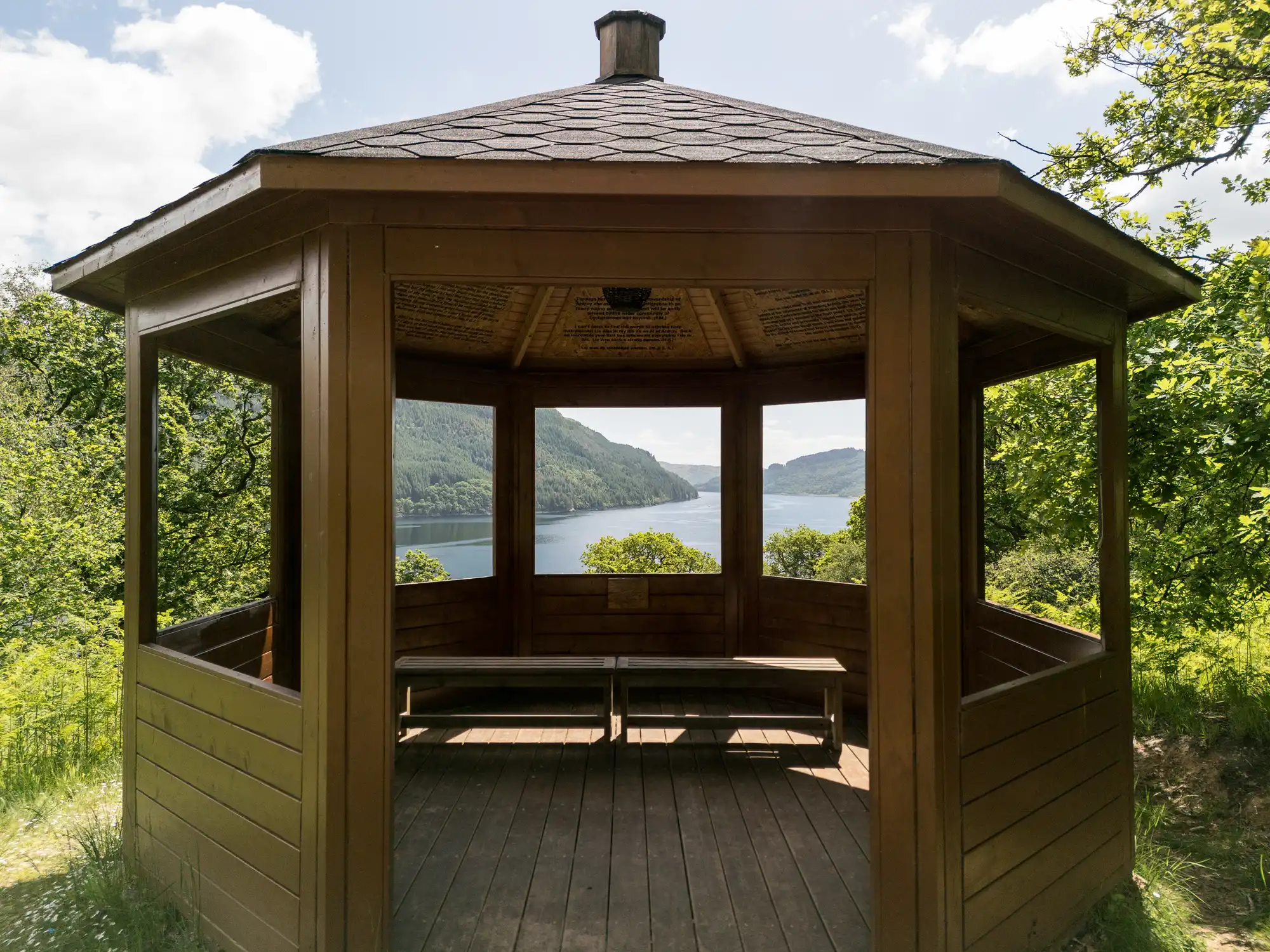 A wooden gazebo with open sides sits on a grassy area overlooking a scenic lake and wooded hills. Sunlight brightens the scene, casting shadows inside the gazebo, where simple benches offer a place to rest and enjoy the natural view.