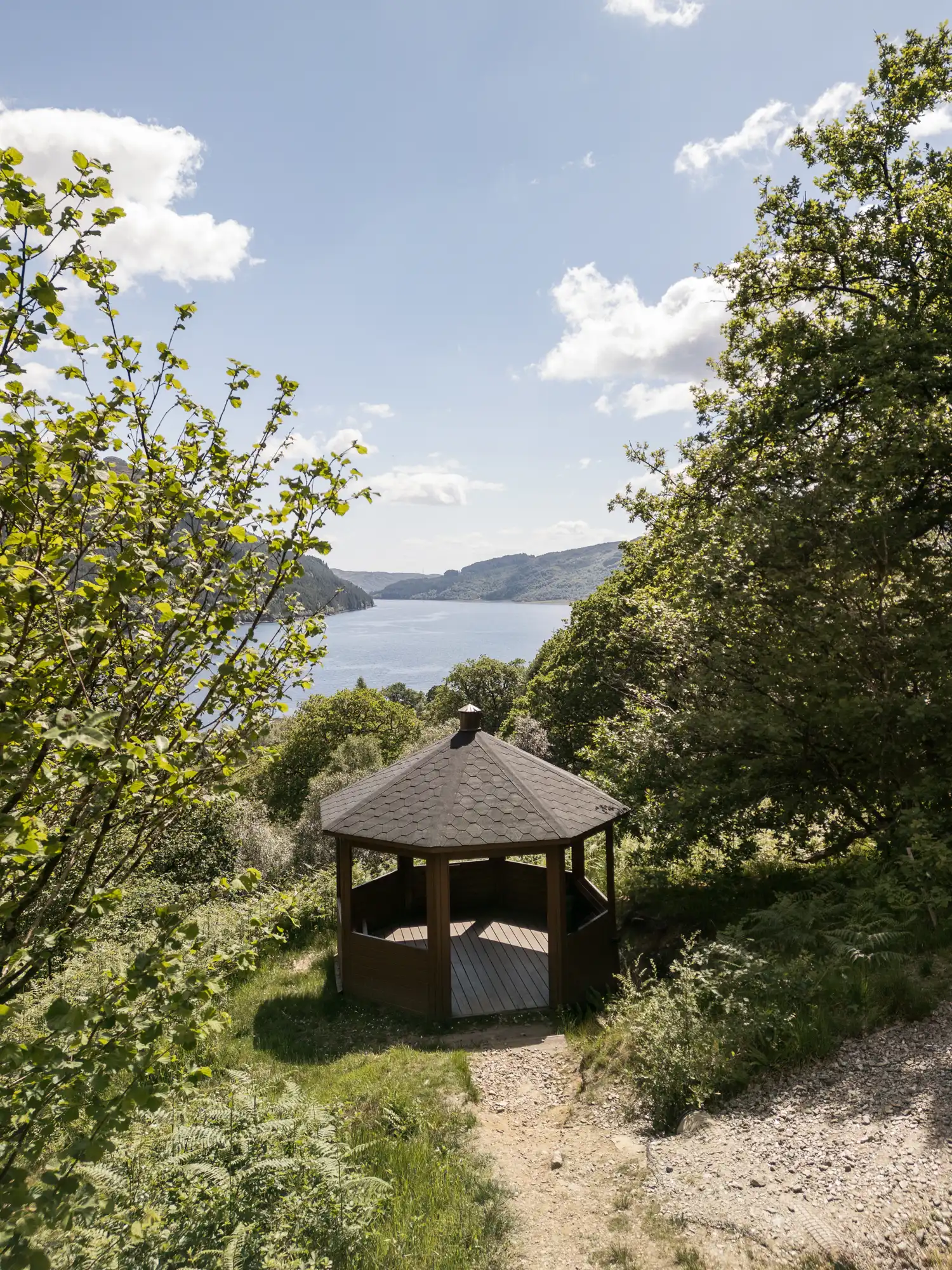 A wooden gazebo with a pointed roof sits in a clearing surrounded by lush green trees, overlooking a lake and distant hills under a partly cloudy blue sky. A narrow dirt path leads to the gazebo through grass and shrubs.