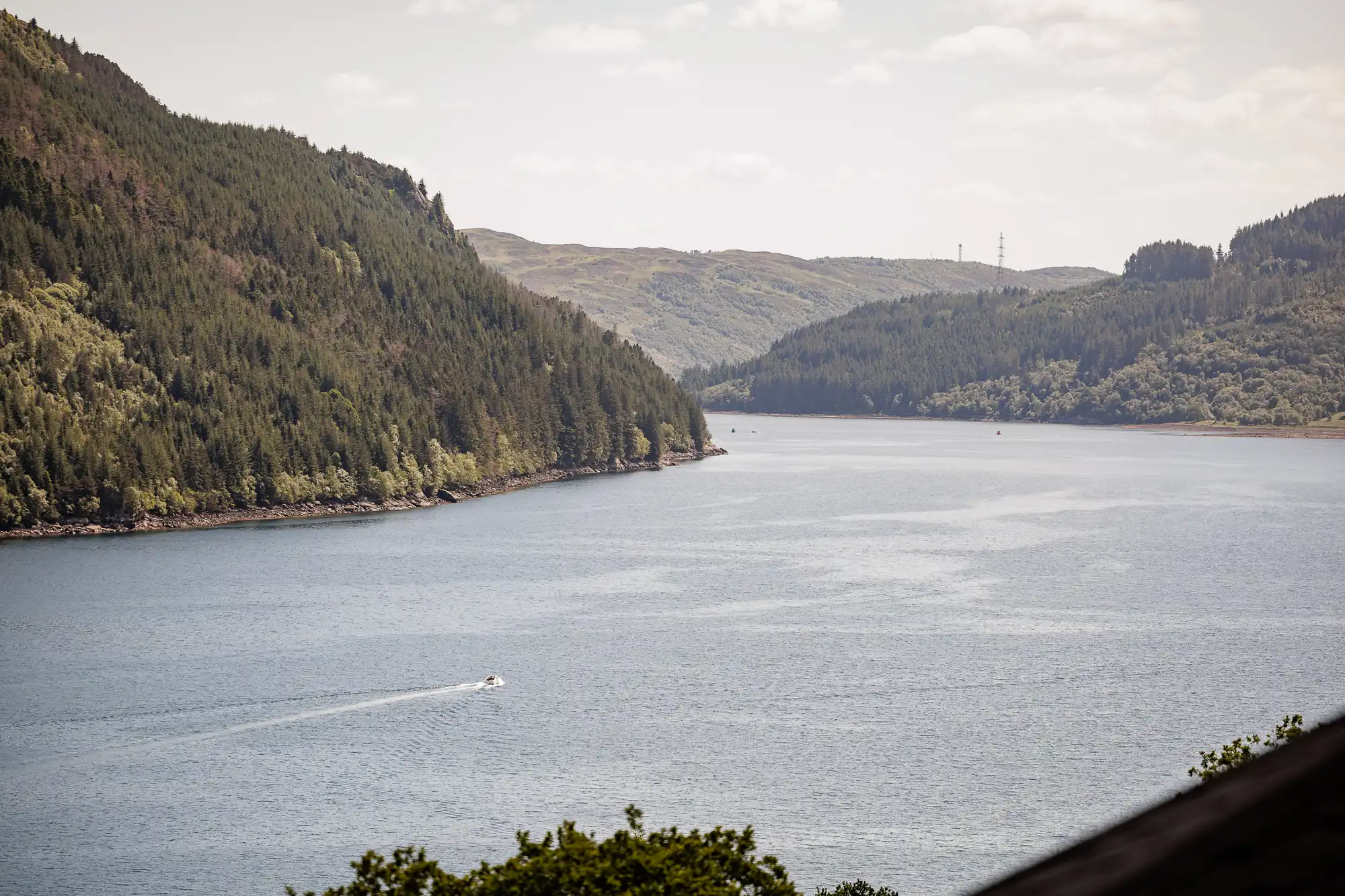 A wide river or lake winds between tree-covered hills under a partly cloudy sky. A small motorboat creates a wake on the calm blue water. Distant hills appear hazy, and the scene feels peaceful and natural.