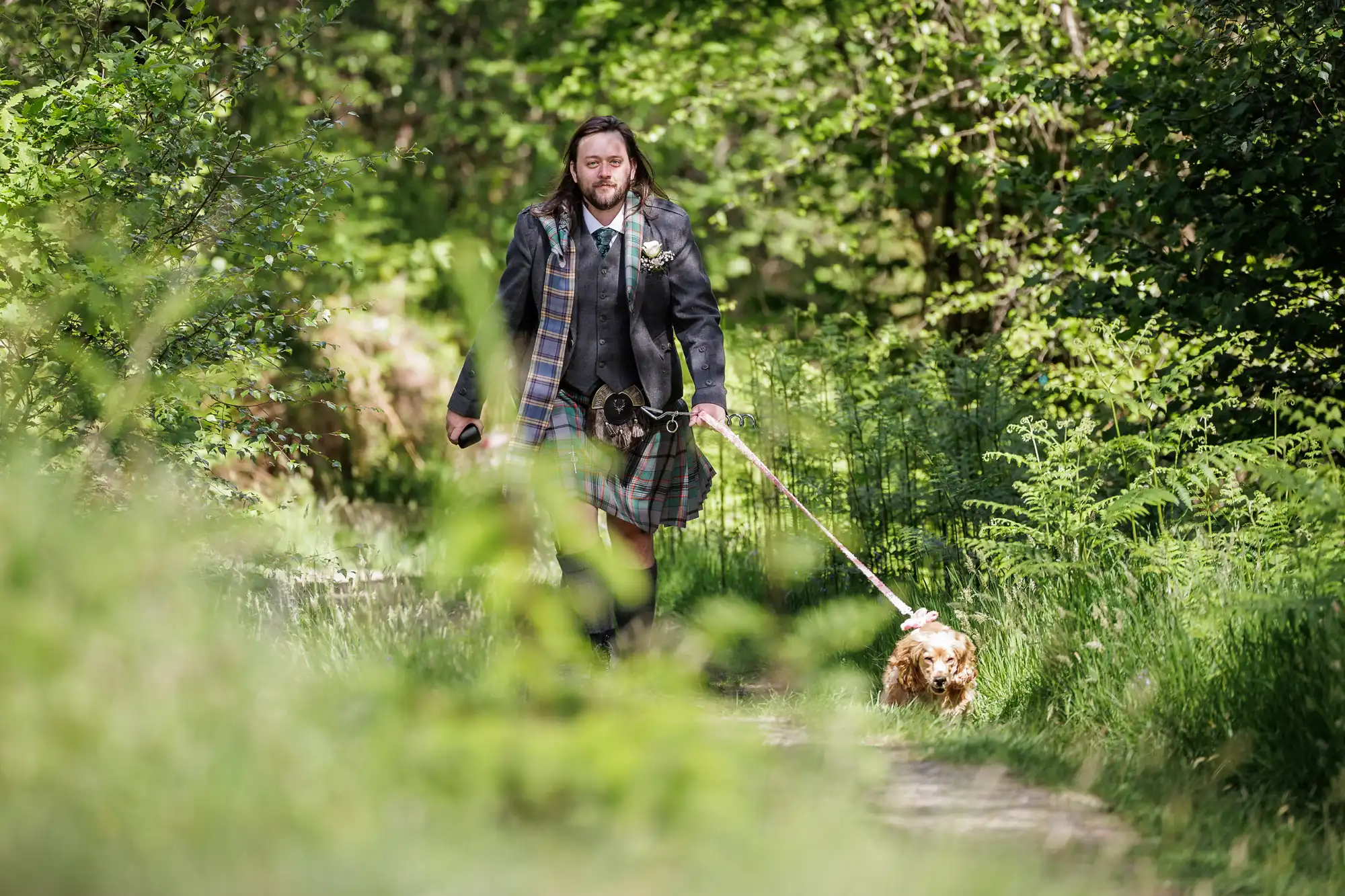 A man in a Scottish kilt and jacket walks a small dog on a lead along a sunlit forest path. Lush green foliage surrounds them, and the man appears relaxed and thoughtful as he strolls through the natural setting.