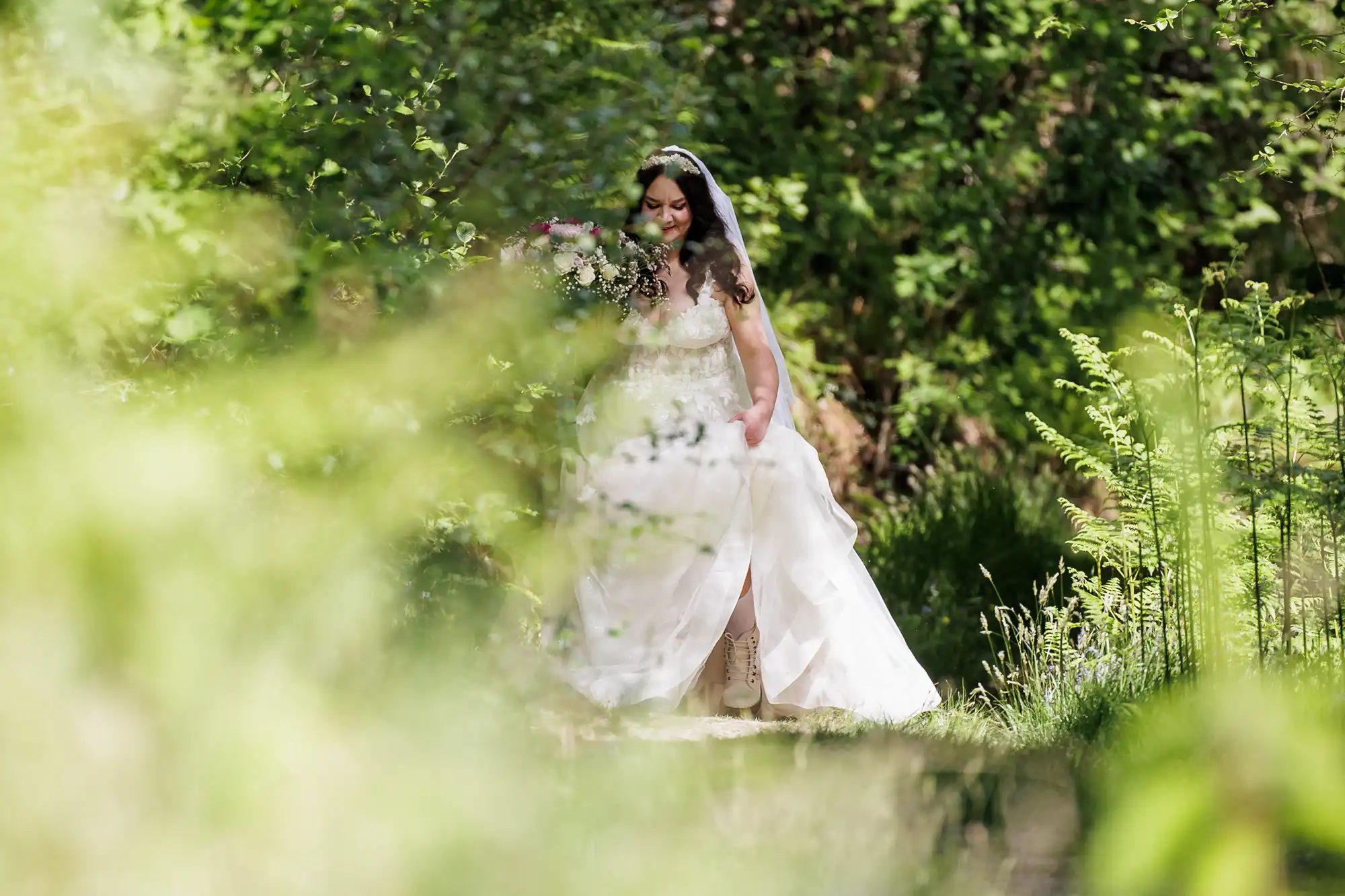 A bride in a white dress and veil walks through a lush, green forest. She holds a bouquet of flowers and looks down, surrounded by sunlight, ferns, and blurred foliage, creating a dreamy, natural atmosphere.