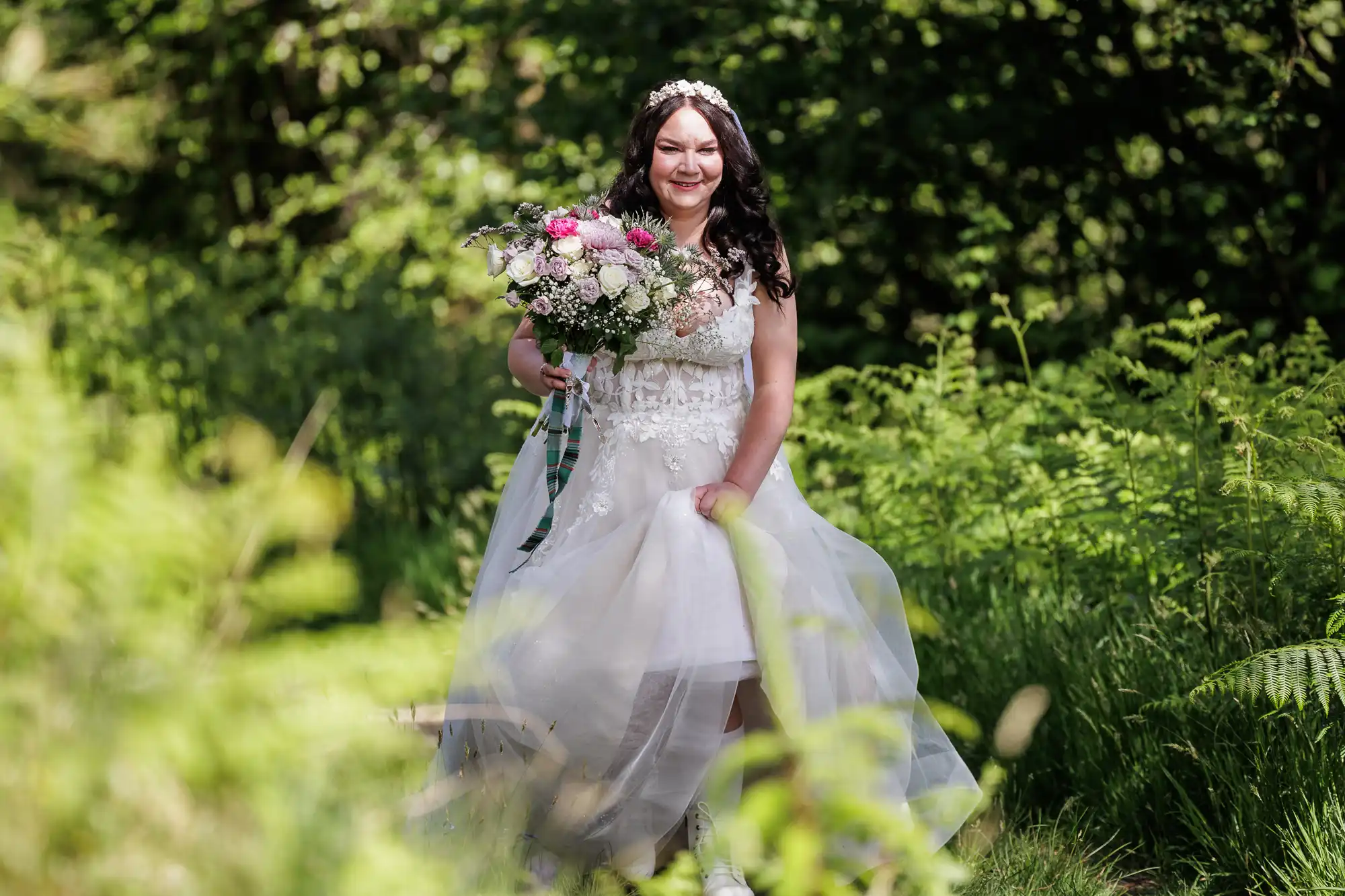 A bride in a white dress and floral headband smiles whilst holding a bouquet of colourful flowers. She walks outdoors on a green, sunlit path surrounded by lush foliage and ferns, with sunlight filtering through the trees in the background.