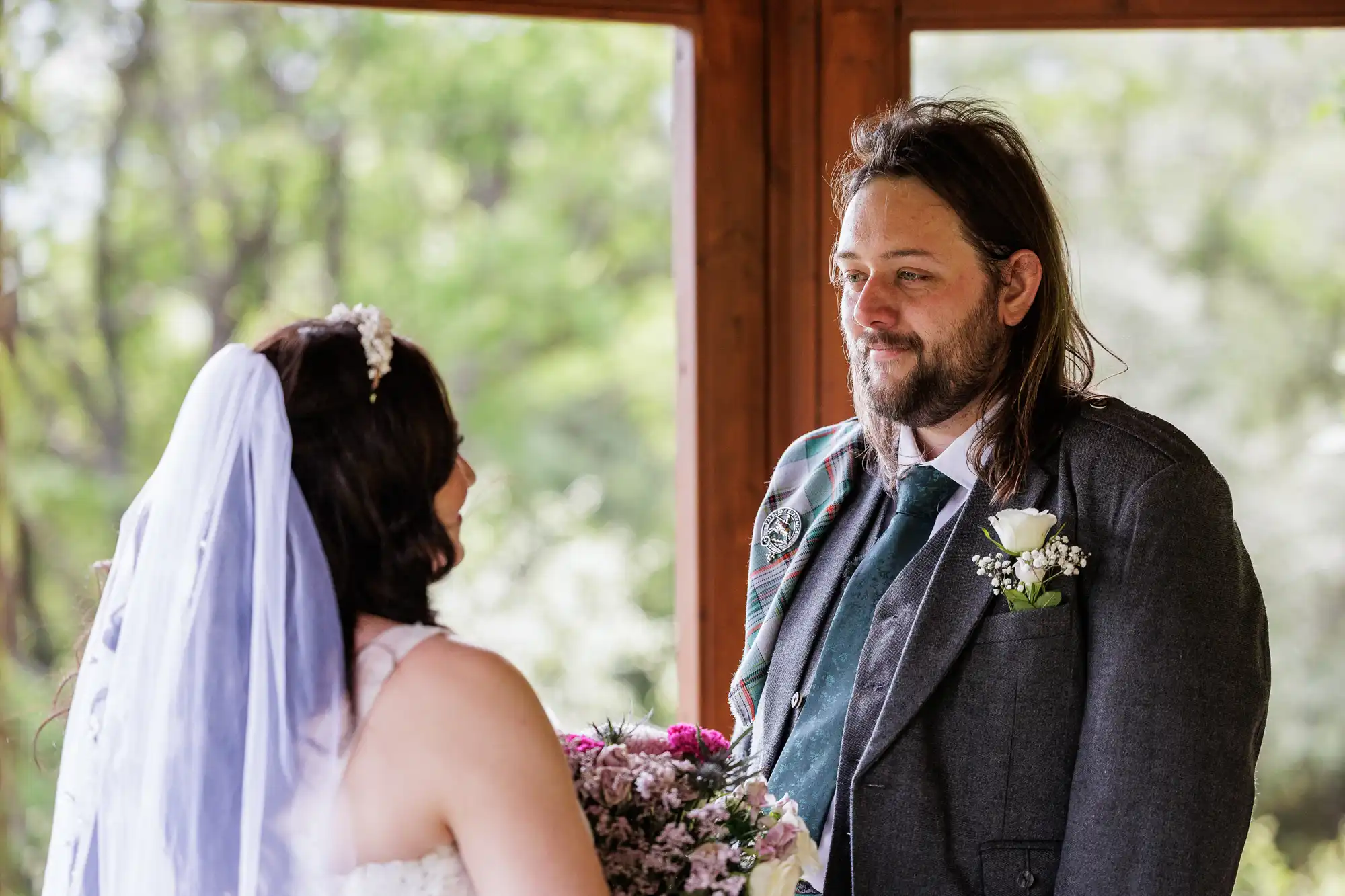 A bride in a white dress and veil holds a bouquet and faces a groom in a grey suit with a checked sash and buttonhole. They stand under a wooden structure, surrounded by greenery, gazing at each other and smiling warmly.
