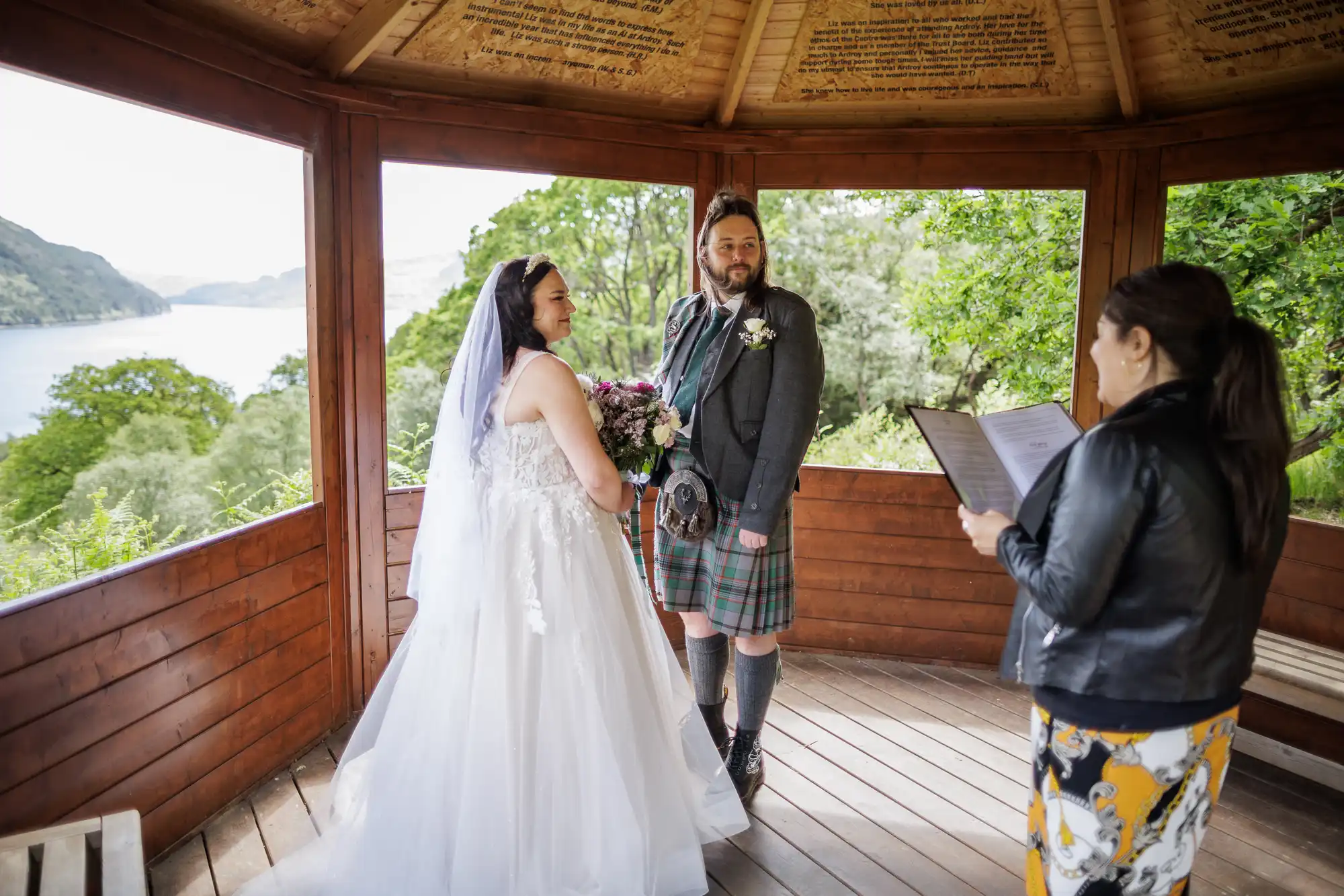 A bride in a white gown and veil stands beside a groom in a kilt inside a wooden gazebo, overlooking a scenic lake and lush greenery. An officiant reads from a folder, conducting the wedding ceremony. Sunlight filters through the open structure.