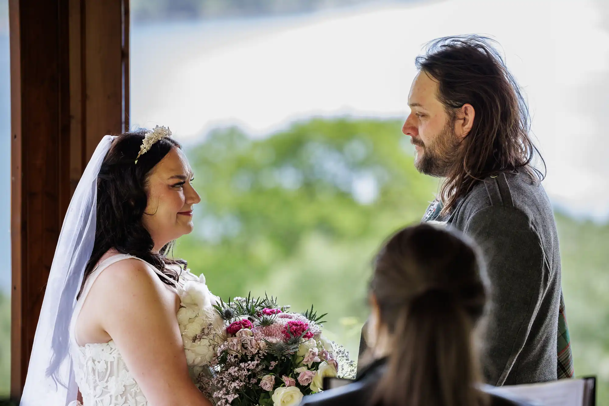 A bride in a white dress and veil holds a bouquet of pink and white flowers, smiling emotionally at a groom with long hair and a beard. They face each other indoors, with a person officiating in the foreground and greenery outside.