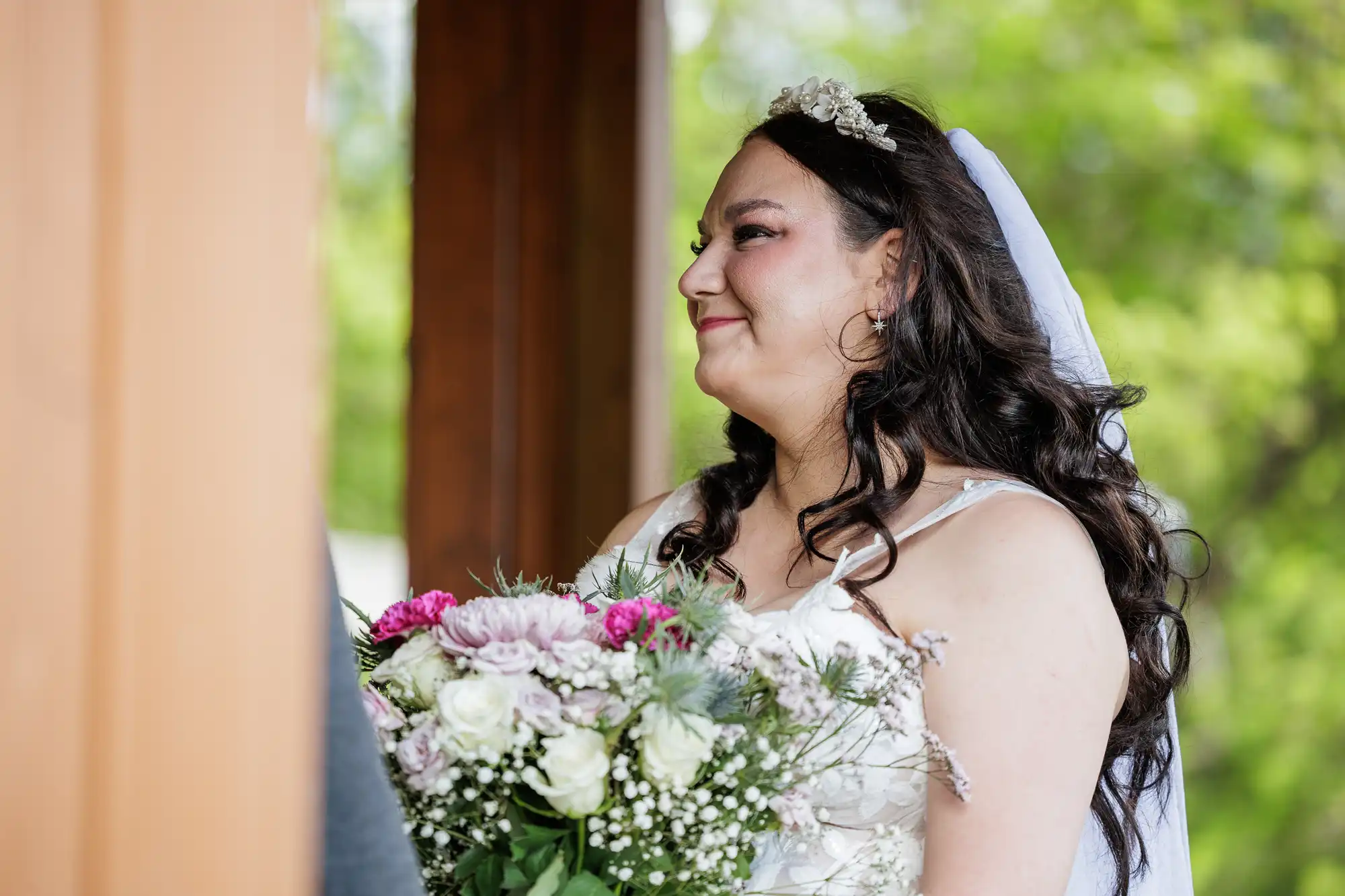 A bride with long dark hair and a jewelled headpiece smiles gently whilst holding a bouquet of white, pink, and purple flowers. She wears a white gown and veil, standing outdoors with greenery blurred in the background.