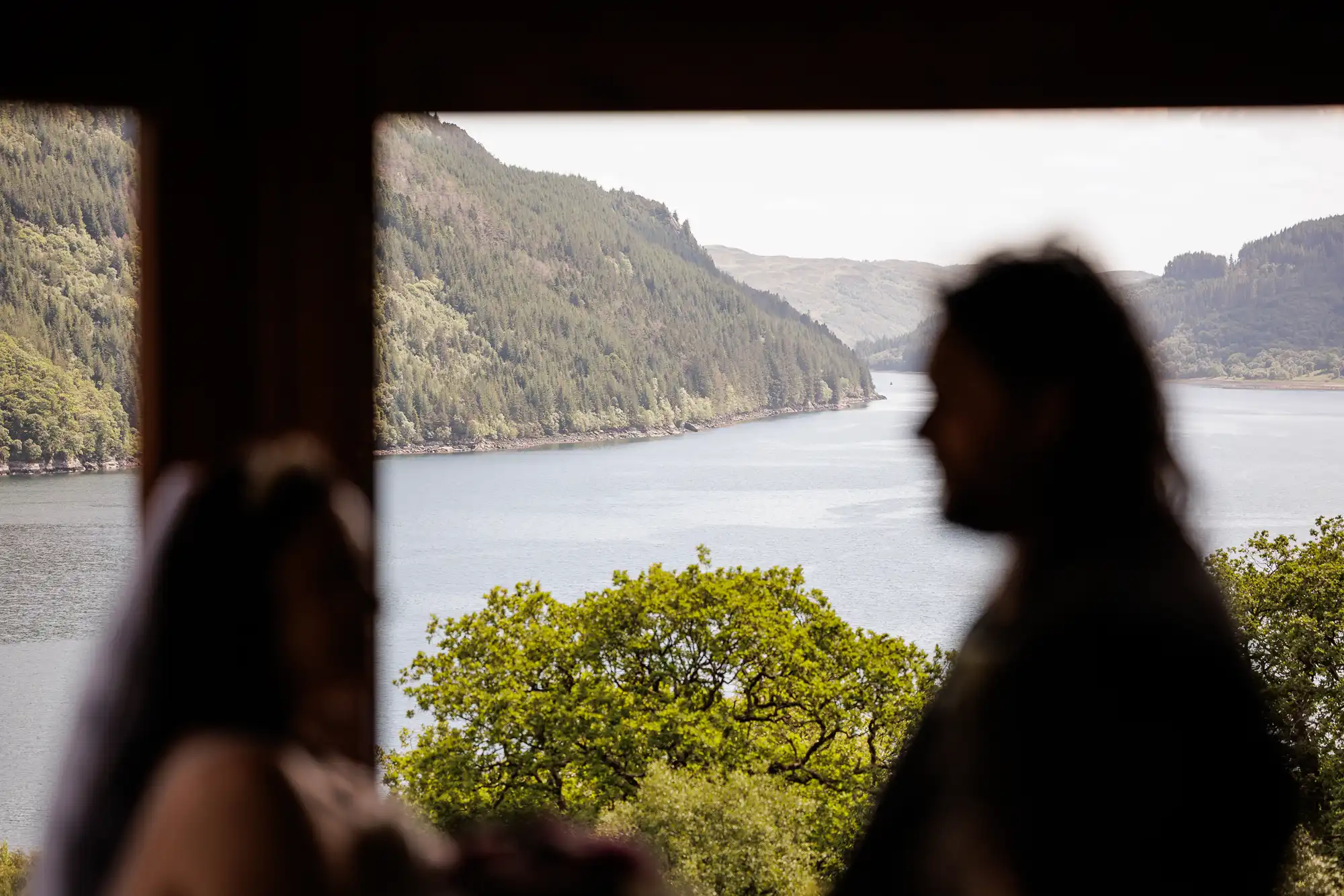 Two people in soft focus, possibly at a wedding, stand in front of a large window. Beyond them is a sharp view of a scenic lake surrounded by green forested hills under a clear sky. The natural landscape is the focal point.