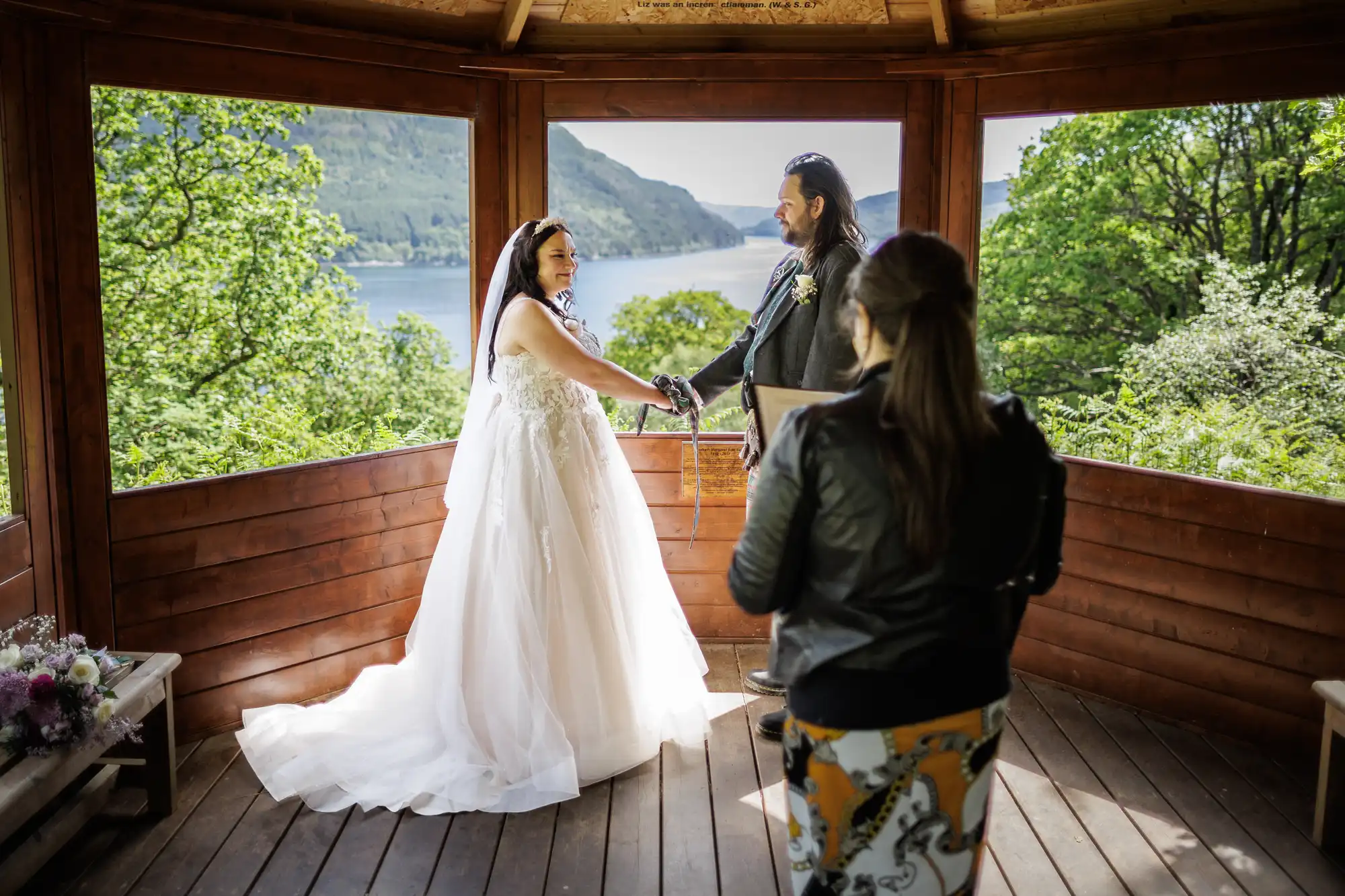 A bride in a white dress and a groom in a dark suit hold hands, smiling, during a wedding ceremony in a wooden gazebo. Lush greenery and a lake are visible outside. An officiant stands with her back to the camera, holding a book.