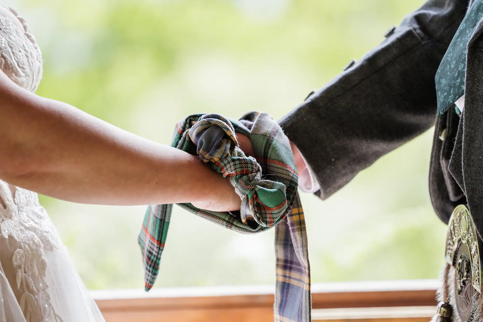 Close-up of two people clasping hands, one in a white lace dress and the other in a dark suit. Their hands are tied together with a knotted tartan cloth, symbolising unity. The background is softly blurred with green foliage.