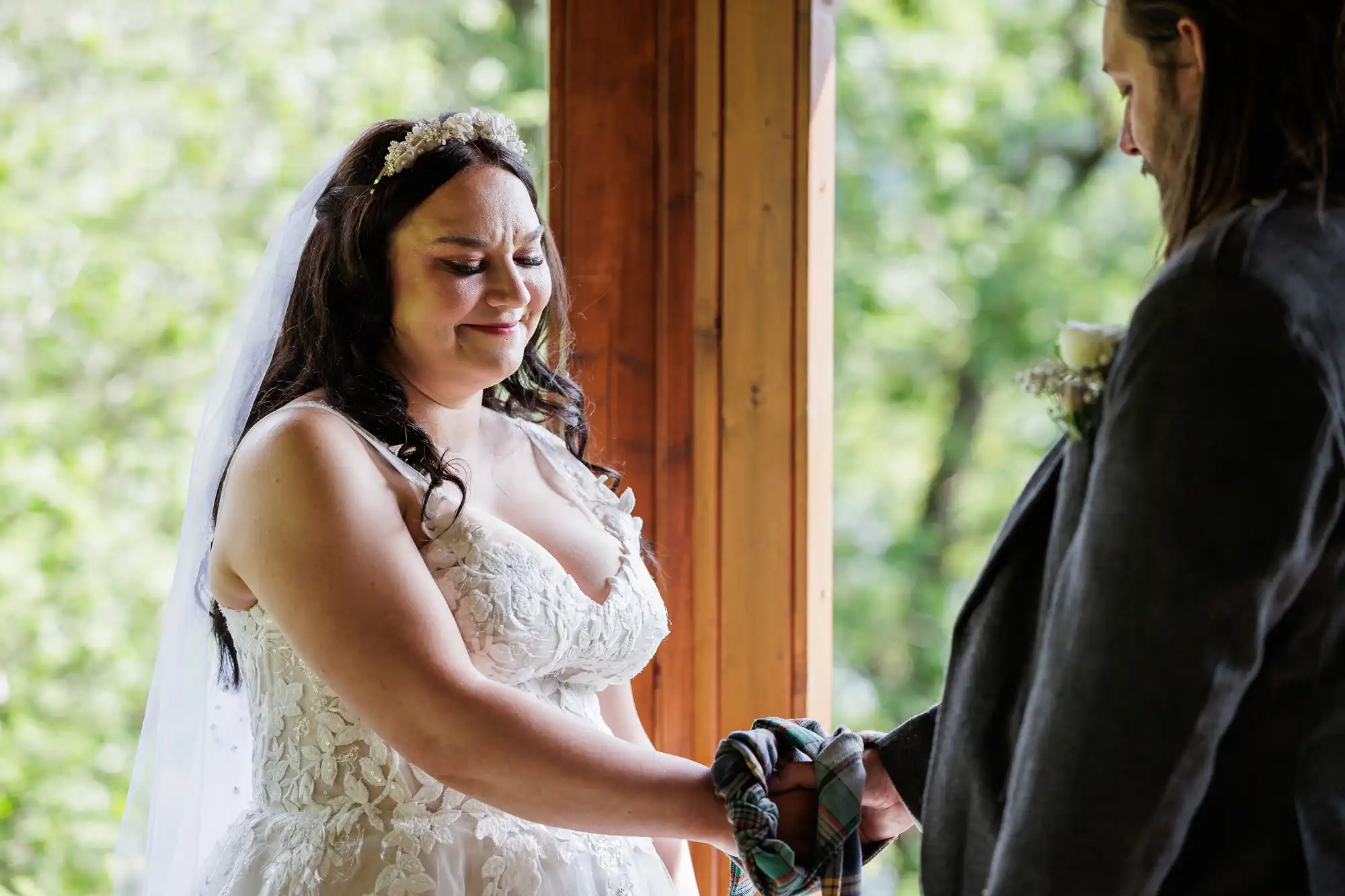 A bride in a white lace dress and veil smiles tearfully as she holds hands with her partner, who wears a dark suit. They stand indoors by a wooden beam, with greenery visible outside. A colourful cloth is wrapped around their joined hands.