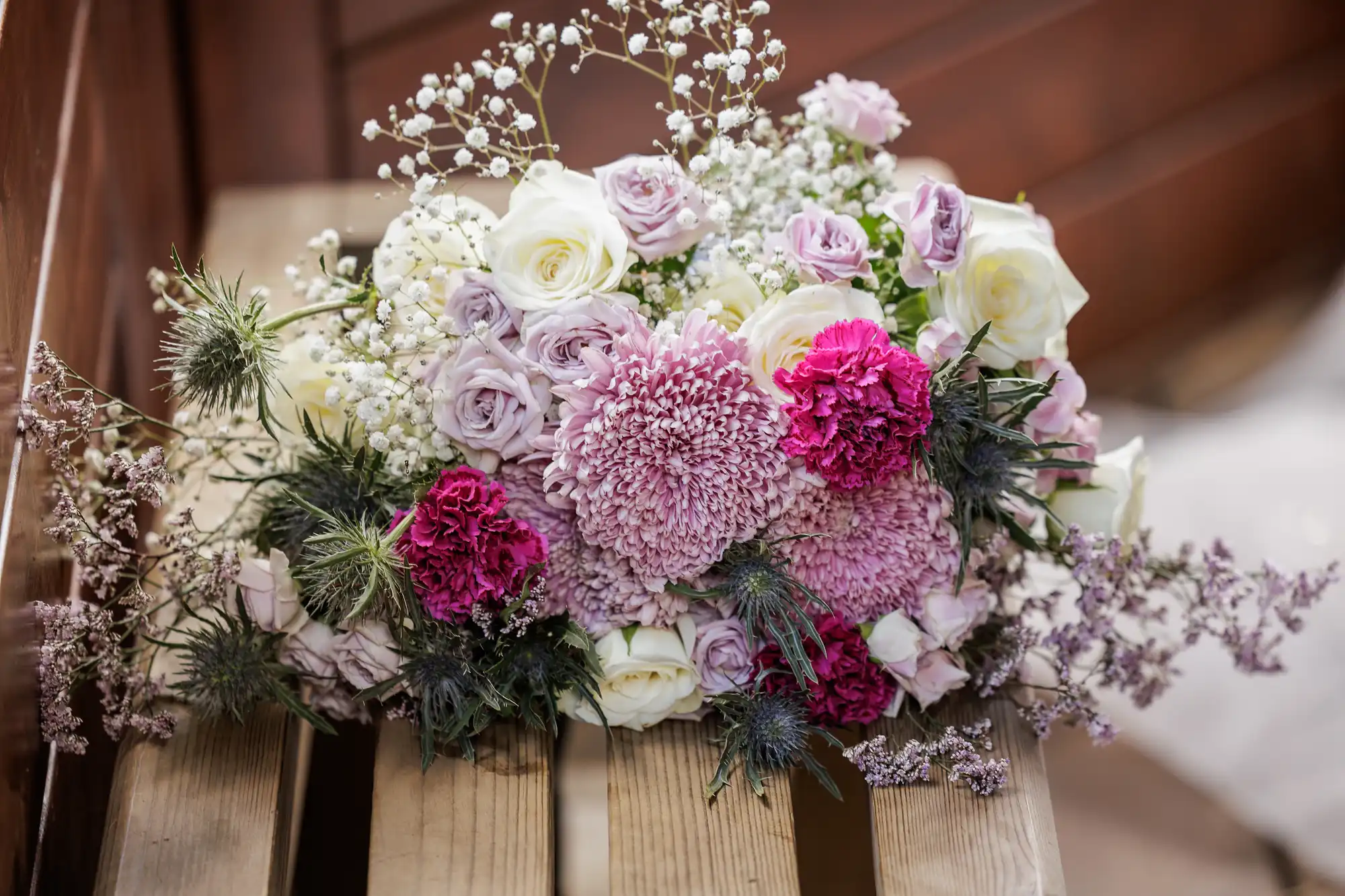 A bouquet of assorted flowers, including white and pale purple roses, pink chrysanthemums, magenta carnations, baby's breath, and greenery, arranged on a wooden bench with a blurred brown background.