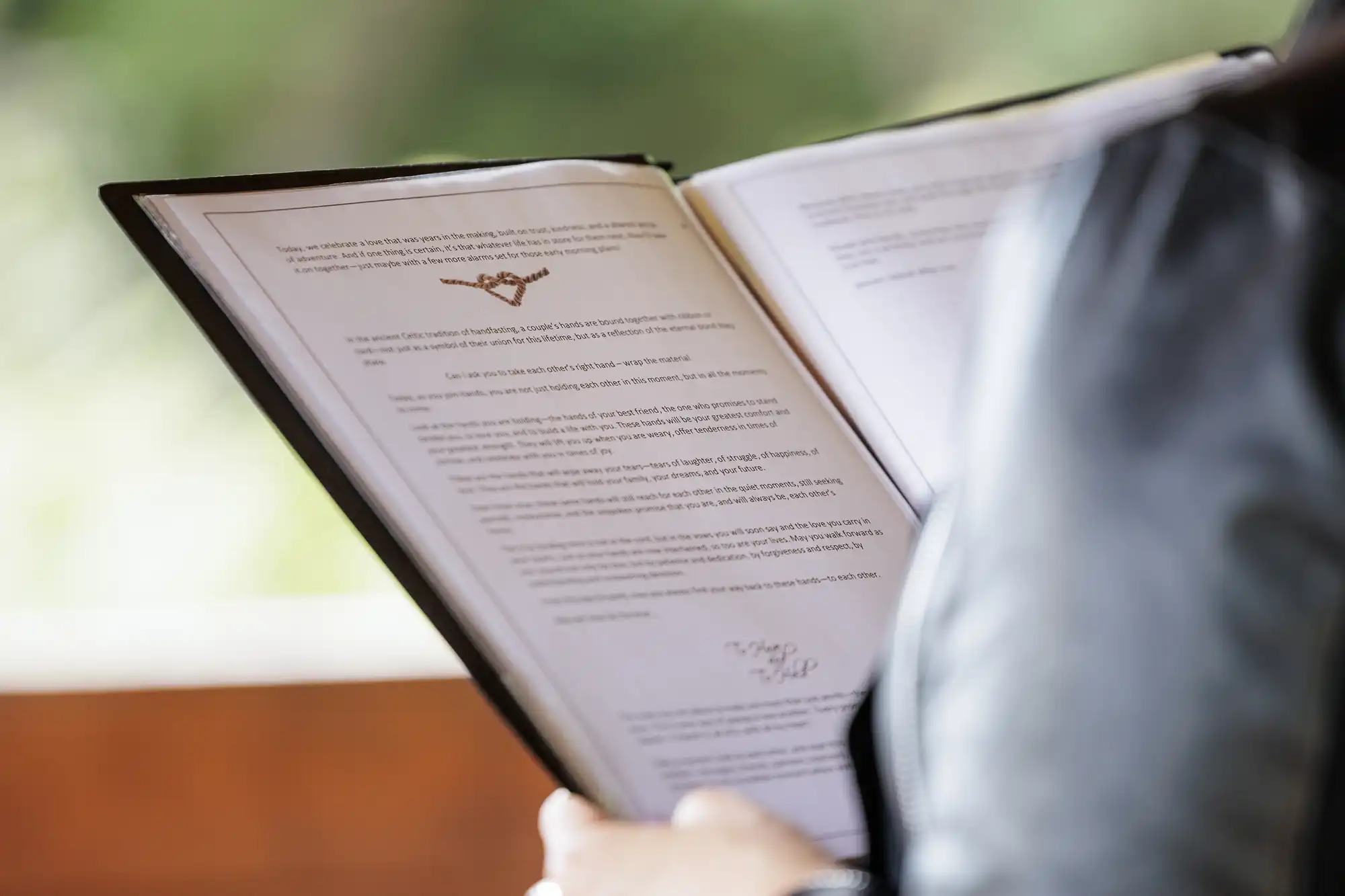 A person wearing a black jacket holds and reads a folder with two typed documents inside, both containing text and signatures. The photo is taken from behind, and the background is blurred greenery and brown, likely outdoors.