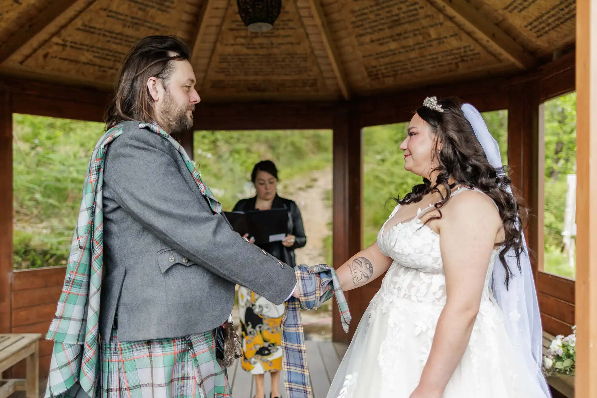 A bride and groom, wearing traditional Scottish attire, stand under a wooden gazebo holding hands with tartan fabric wrapped around them. The bride smiles in a white dress and veil; an officiant stands behind them reading from a folder.