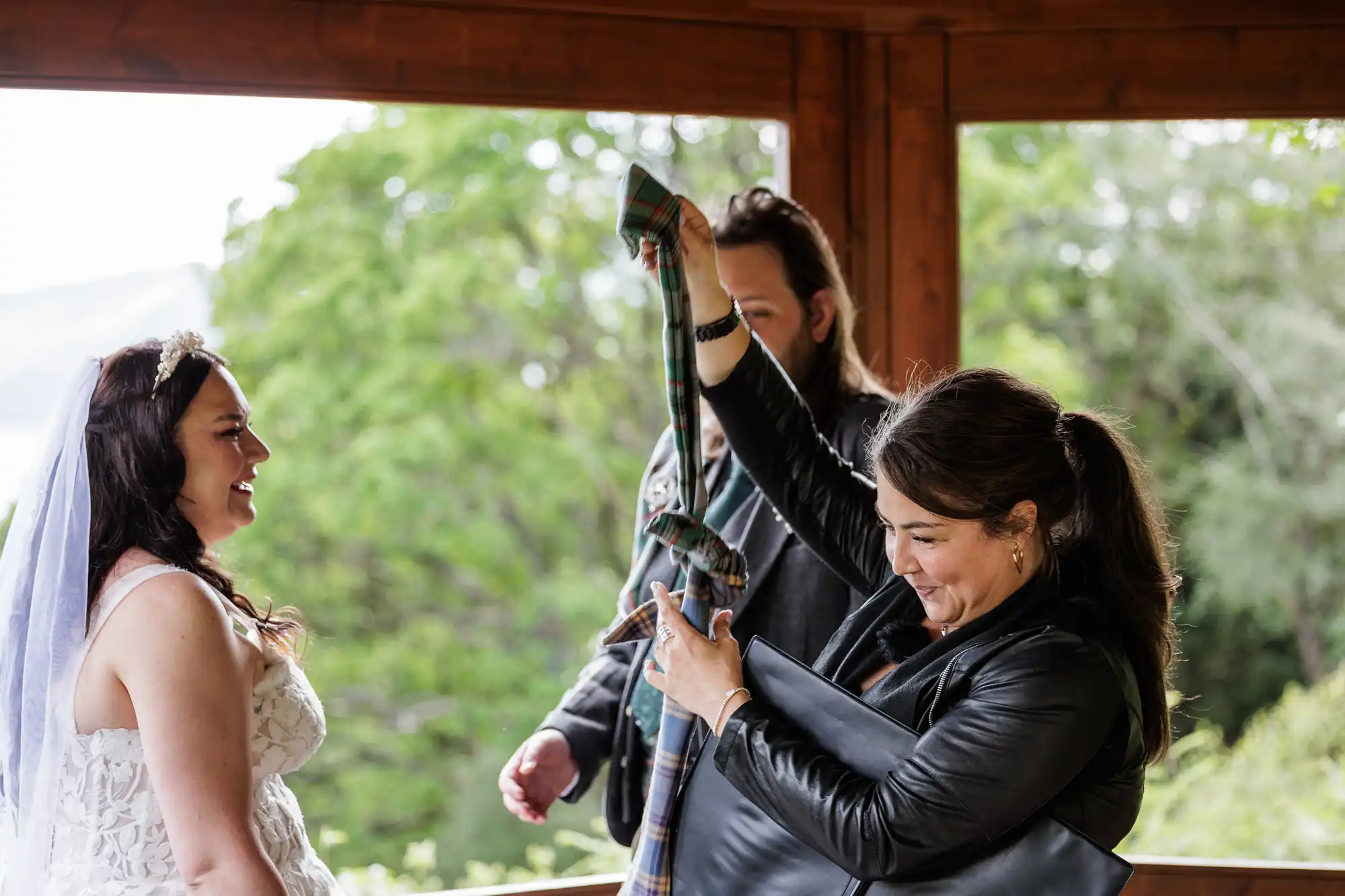 A woman in a white wedding dress and veil smiles at two people. One woman, dressed in black, holds up a long, patterned cloth with joy, whilst a man stands behind her. They are under a wooden structure with greenery in the background.