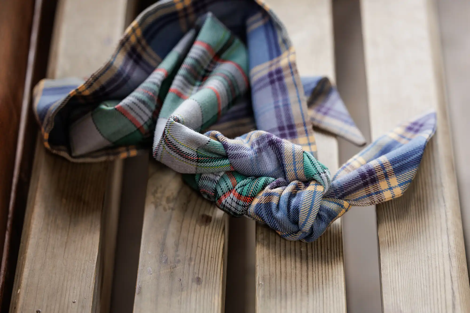 A checked fabric hairband with knotted ends lies on a wooden slatted bench. The fabric features various patterns in blue, green, yellow, and red, creating a casual and cosy look. The bench has a weathered, rustic appearance.