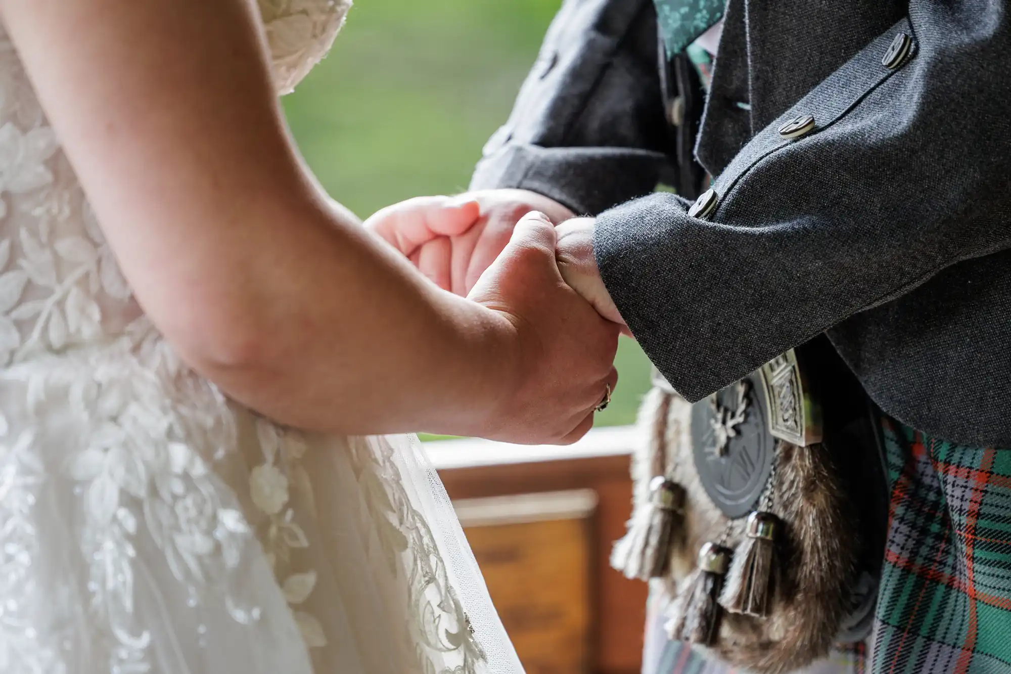 A close-up of a couple holding hands at a wedding. The bride wears a white lace dress, whilst the groom wears a traditional Scottish outfit with a tartan kilt and sporran. Their hands are gently clasped, symbolising love and commitment.