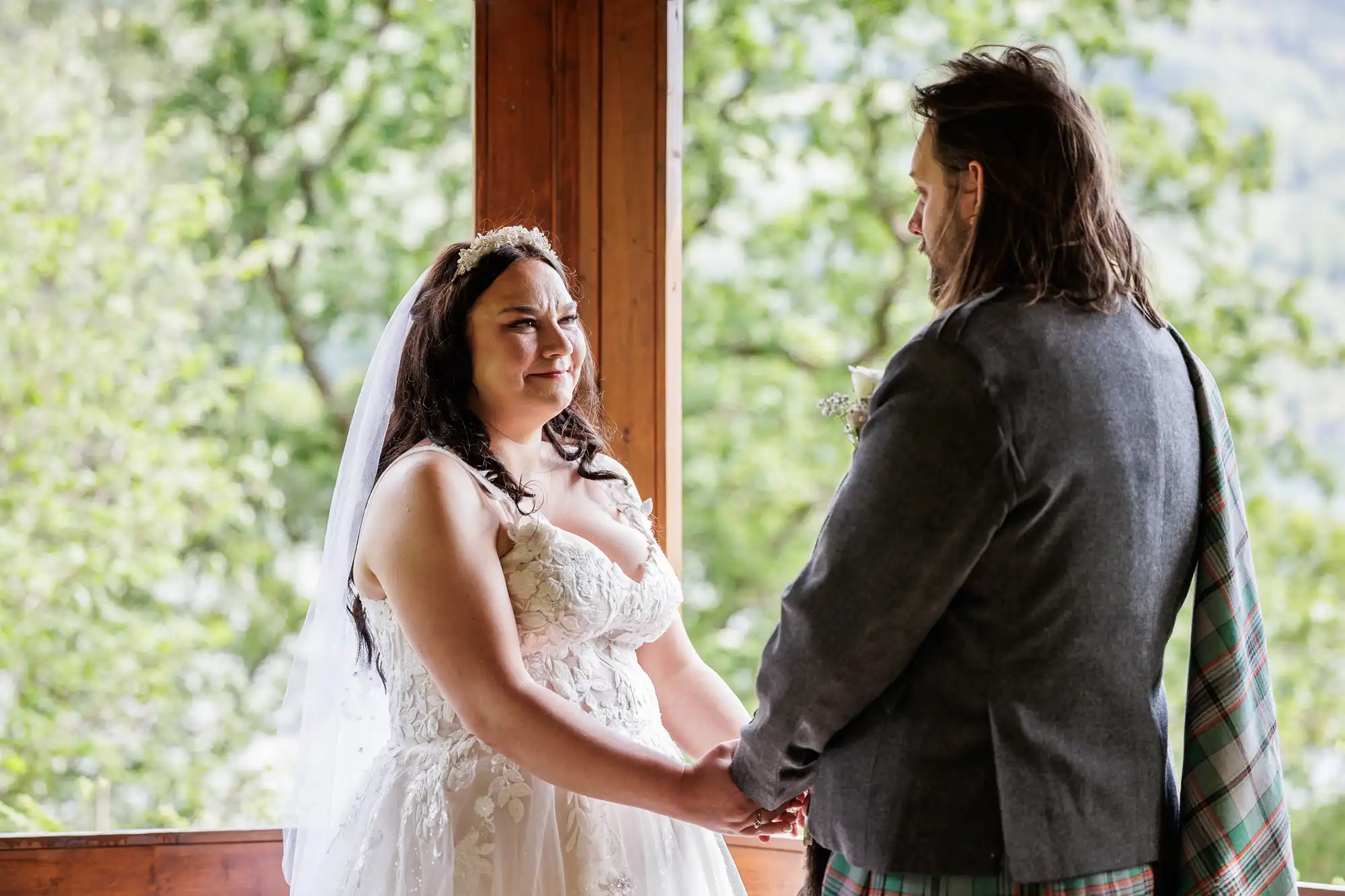 A bride in a white lace dress and veil holds hands with a groom in a grey suit and tartan sash. They face each other, standing on a wooden veranda with green trees and natural light in the background. The bride appears emotional and is smiling.