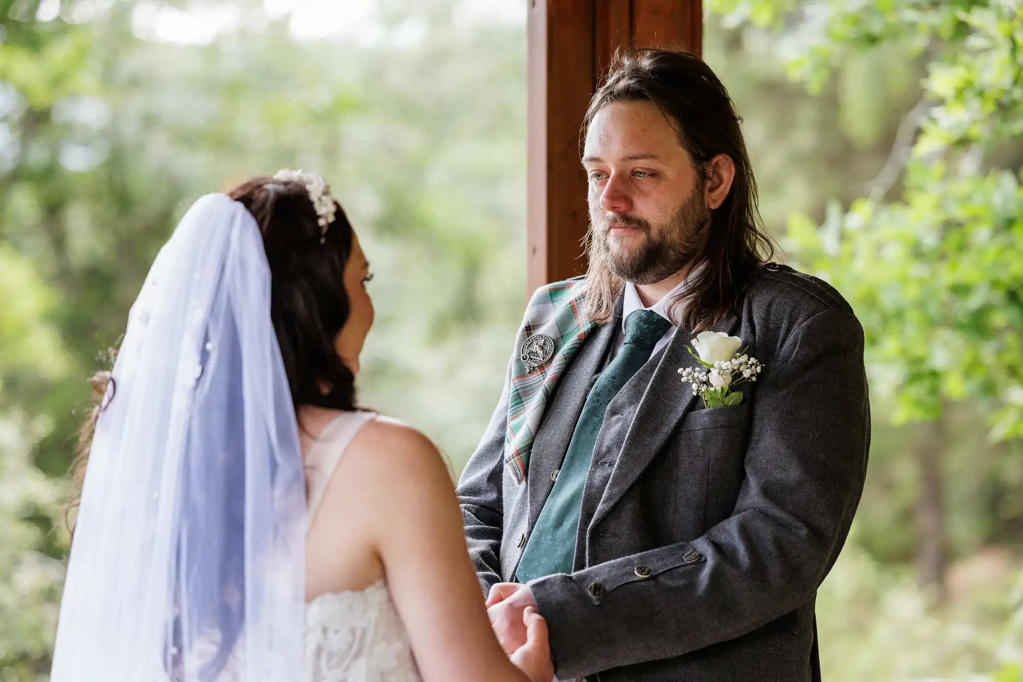A bride in a white dress and veil holds hands with a groom in a grey suit with a tartan sash and green tie. They face each other under a wooden structure, surrounded by lush green trees, sharing an emotional, intimate moment.