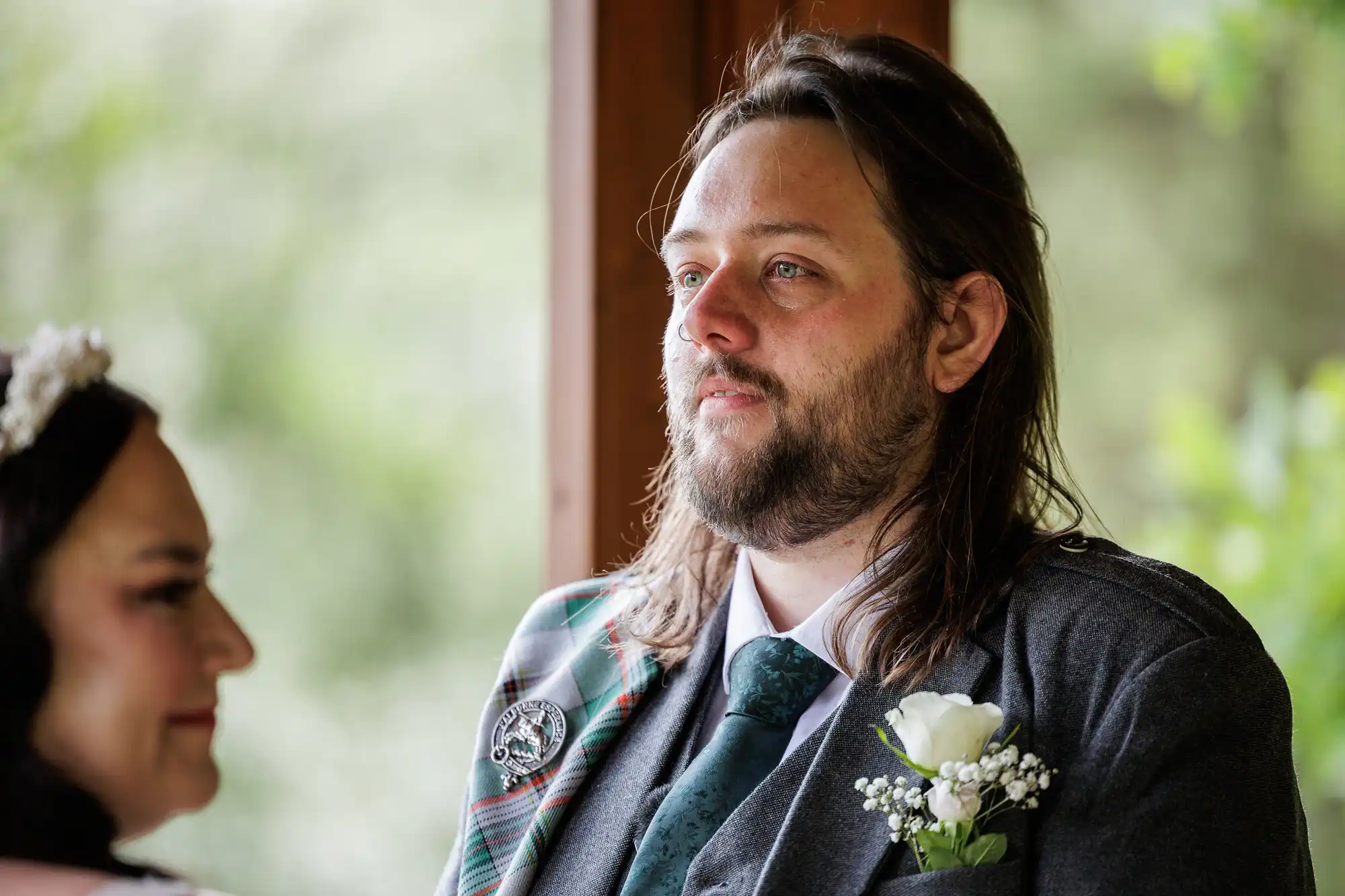 A man with long brown hair and a beard, dressed in a grey suit, checked sash, and green tie, looks emotional. He has a white rose buttonhole. A woman in a white dress and tiara is partially visible beside him. The background is softly blurred greenery.