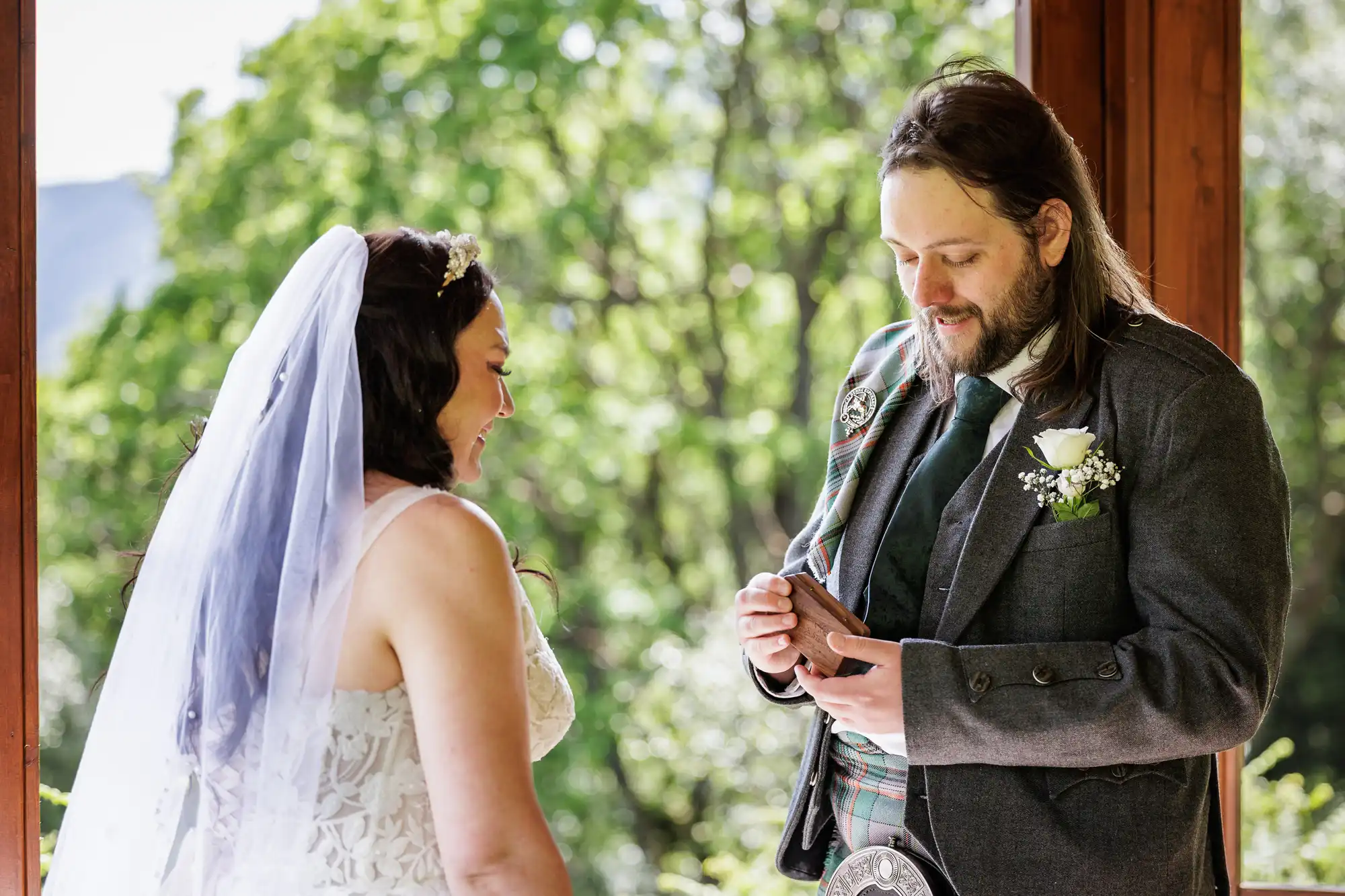 A bride in a white dress and veil smiles as a groom in a kilt and suit jacket places a ring on her finger. They stand in front of a window with lush green trees outside, sunlight illuminating the scene.
