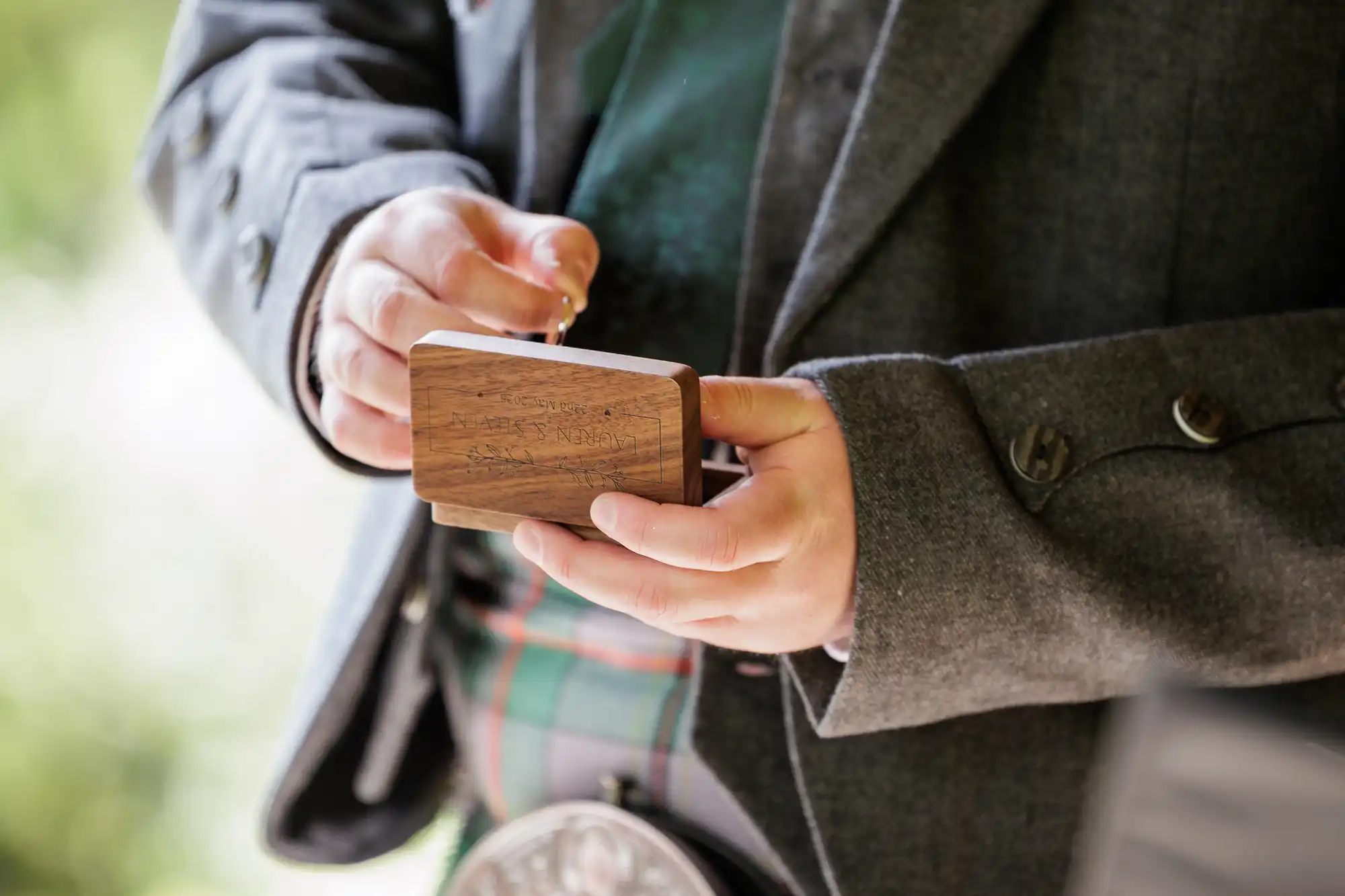 A person in a grey jacket and tartan kilt holds a small wooden box with engraved text. The focus is on their hands and the box, whilst the background is blurred with green hues. Part of a traditional sporran is visible at the waist.