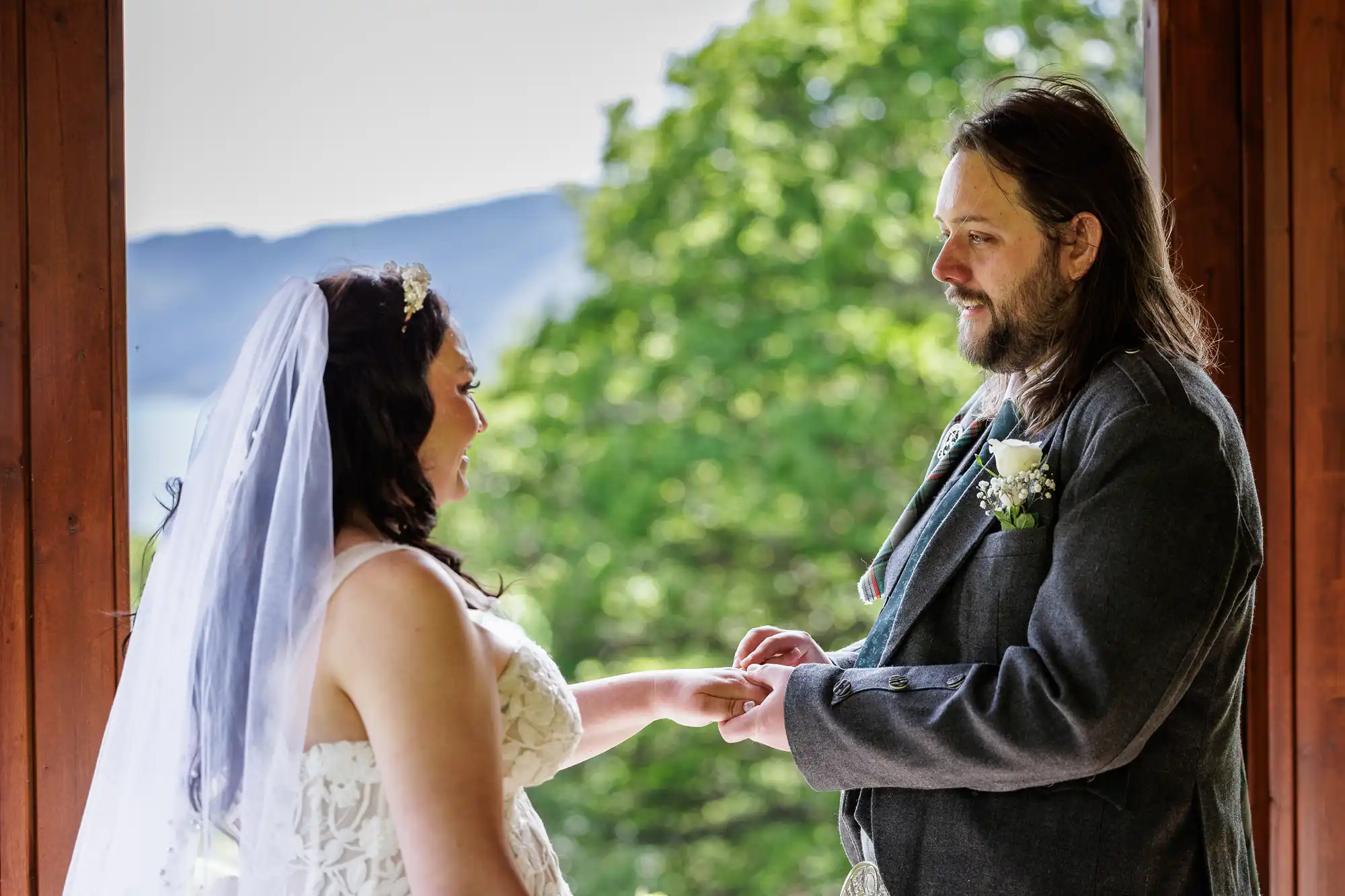 A bride in a white dress and veil smiles as a groom in a grey suit and kilt gently holds her hand, placing a ring on her finger. They stand in front of a wooden frame with lush green trees and distant hills in the background.